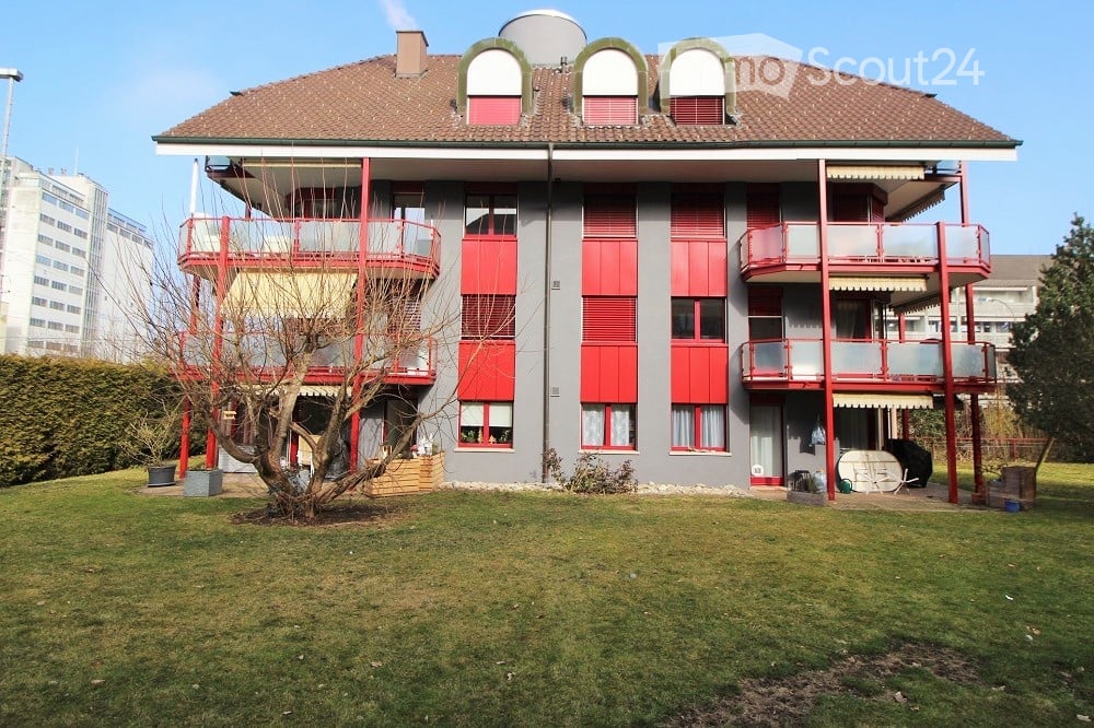 Multi-story apartment building with red shutters, balconies, and a well-maintained lawn in front.