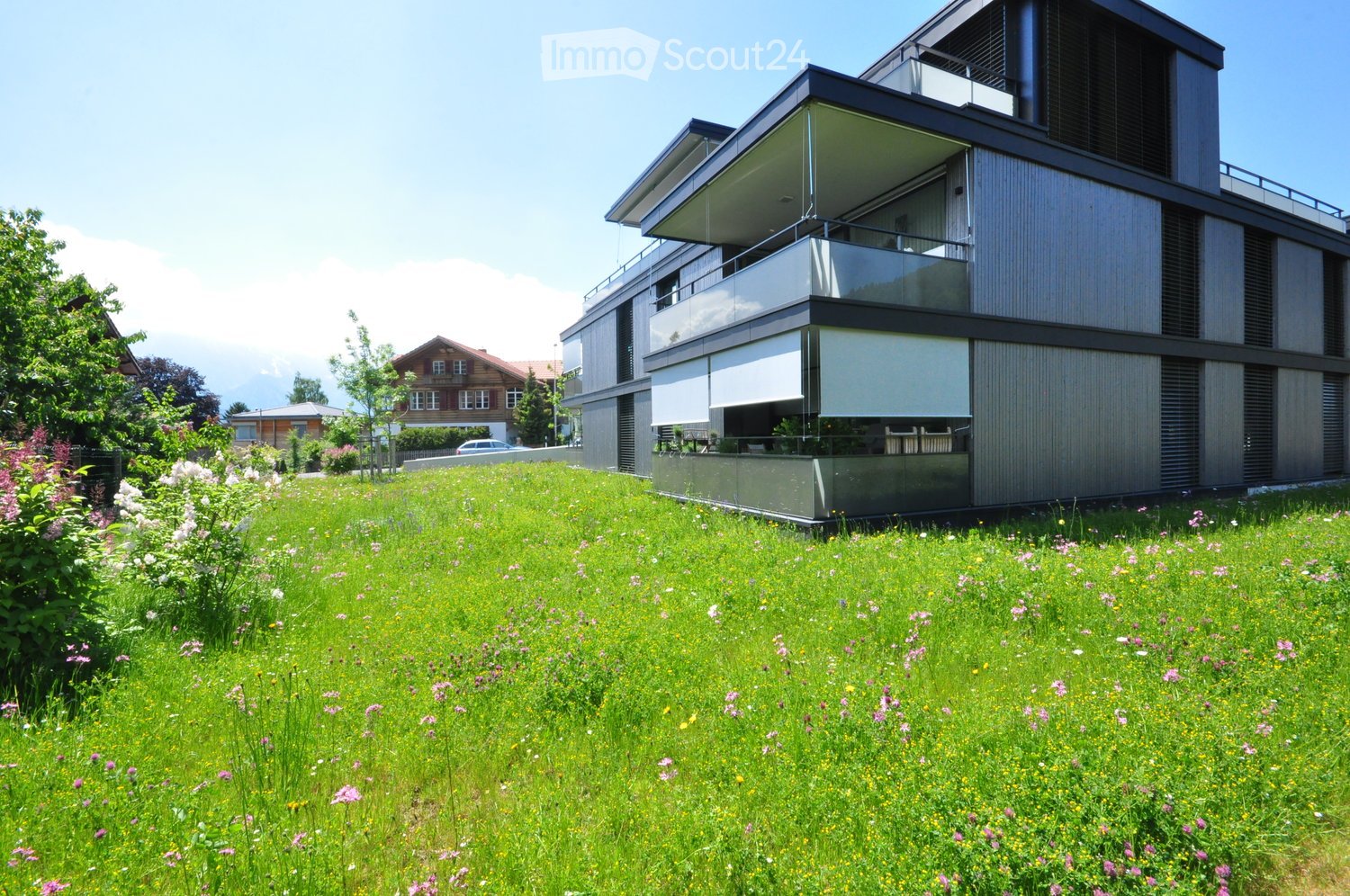 Modern two-story house with a raised ground floor, large windows, and a balcony. The surrounding area features a well-maintained lawn with colorful flowers and shrubs.