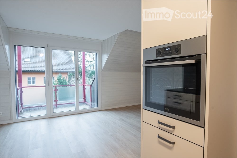 Bright, unfurnished room with wooden flooring, large sliding glass doors leading to a balcony, and a modern kitchen area with a built-in oven.
