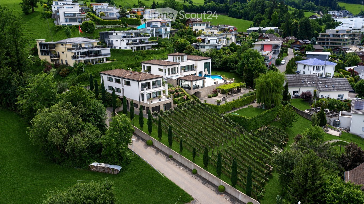 Aerial view of a residential area with multiple modern houses, green lawns, trees, and a swimming pool visible in the foreground.