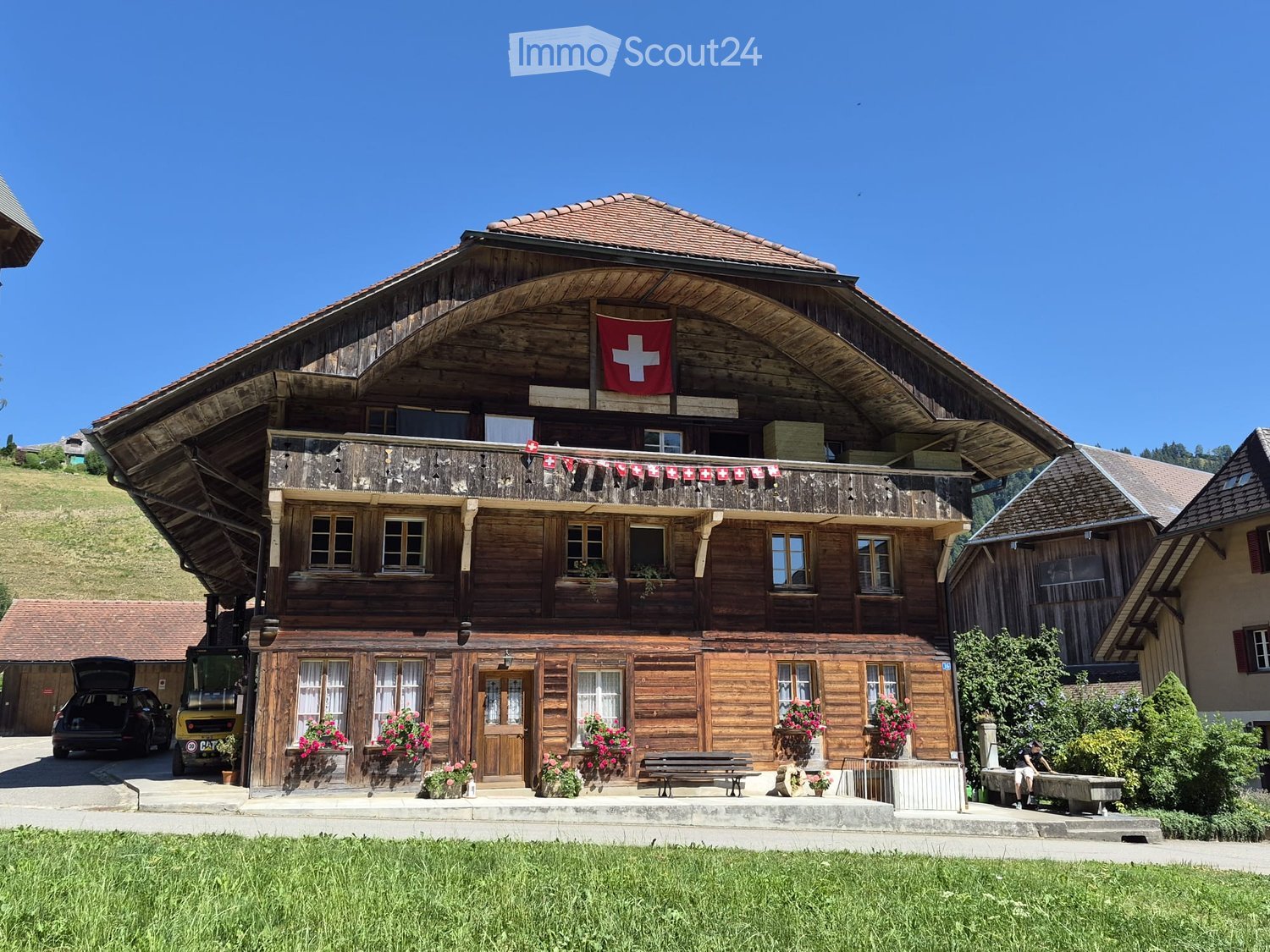 Traditional wooden house with a large balcony, multiple windows, and Swiss flags, surrounded by greenery and a parked vehicle.