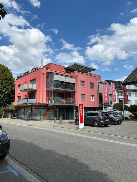 Modern red building with multiple floors, balconies, and a storefront on the ground level. Several cars are parked in front.