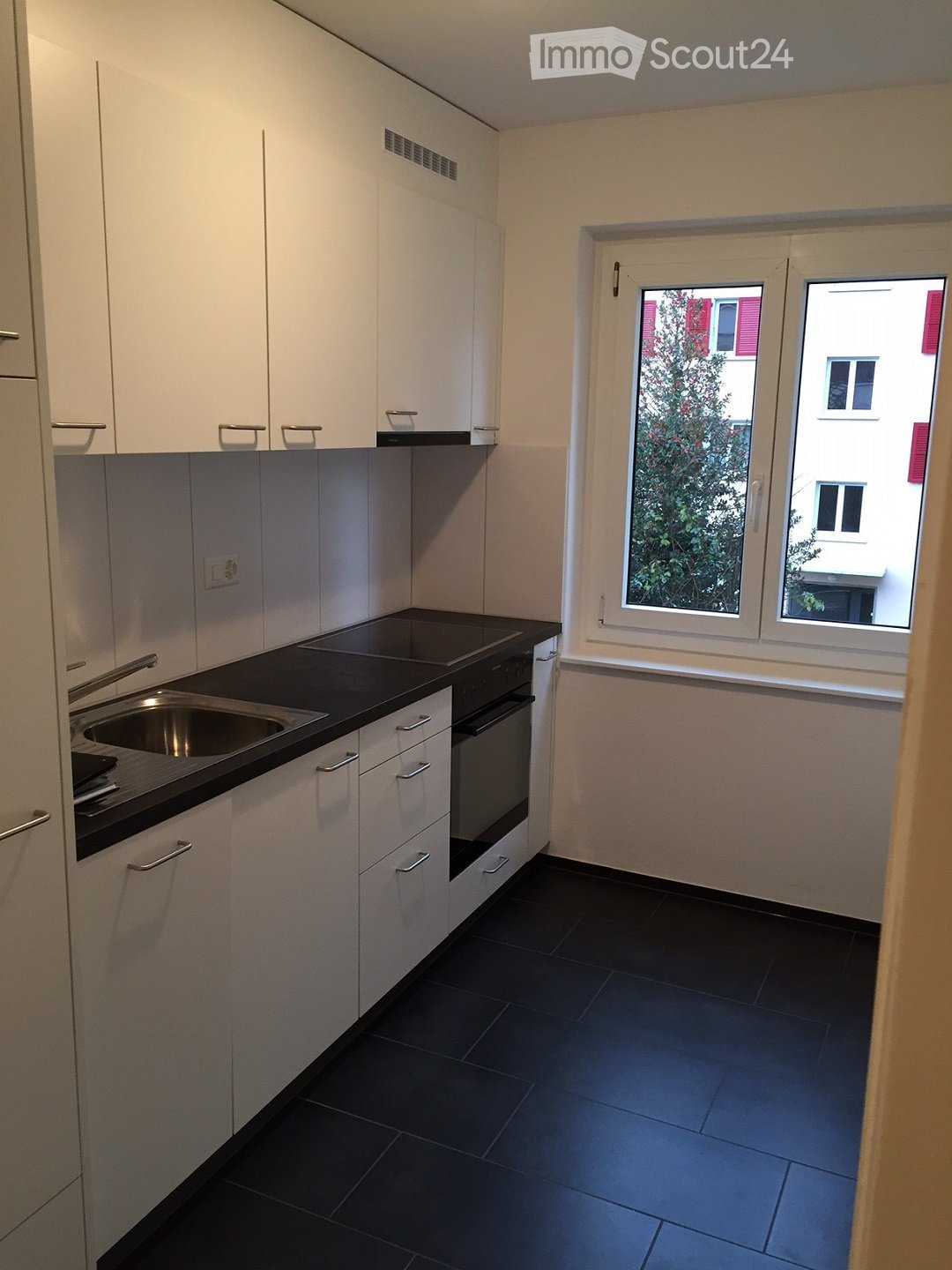 Modern kitchen with white cabinets, black countertop, stainless steel sink, and built-in oven. The space features dark tiled flooring and large windows offering a view of greenery and neighboring buildings.