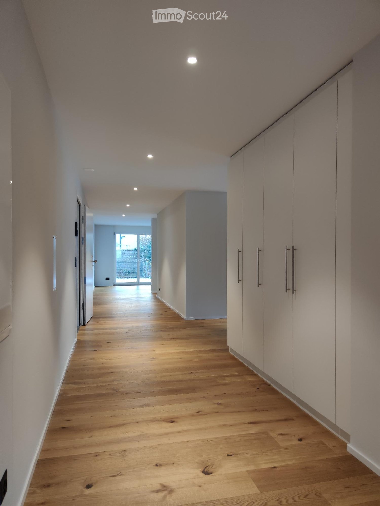 Modern hallway with light wooden flooring, white walls, built-in storage units, and recessed lighting.