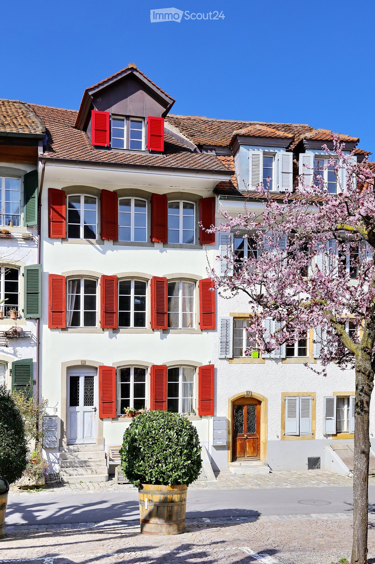 A two-story white house with red shutters, multiple windows, and a small garden area with a tree and a barrel planter.