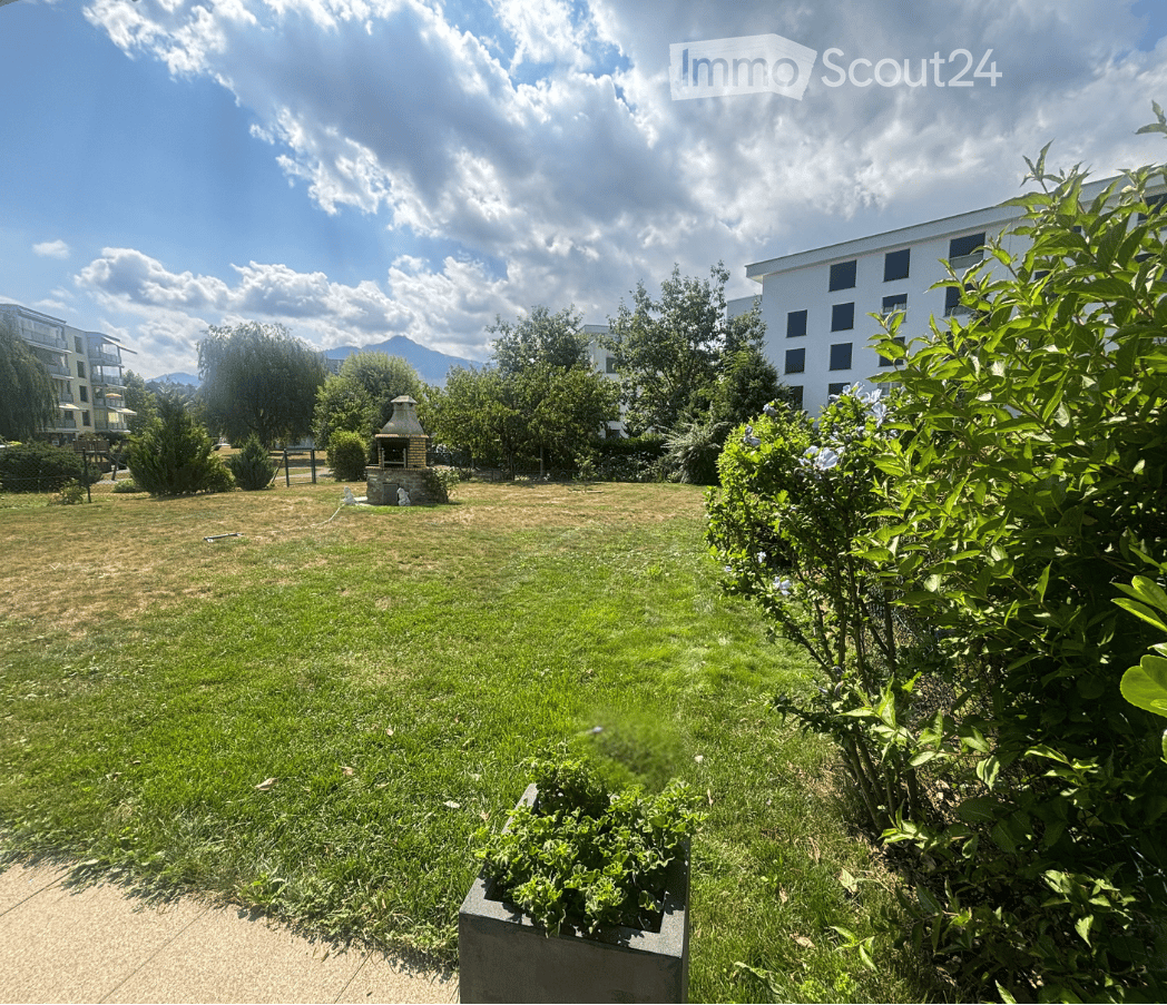 A spacious, well-maintained garden area with green grass, trees, shrubs, and a small stone fireplace in the background. The area is bordered by a modern building and features a clear, partly cloudy sky.