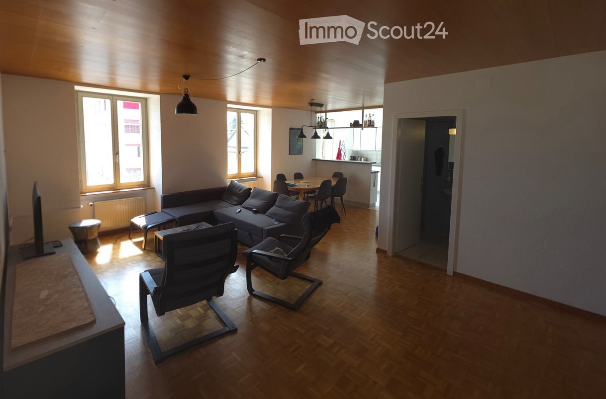 Bright living room with wooden flooring, large windows, a sectional sofa, chairs, a coffee table, and a TV stand.