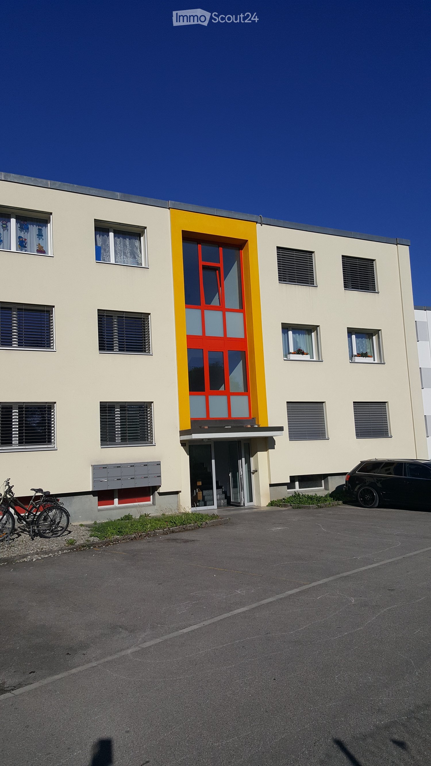 Two-story building with light beige walls, red and yellow entrance door, multiple windows with blinds, and a small parking area with bicycles and a car.