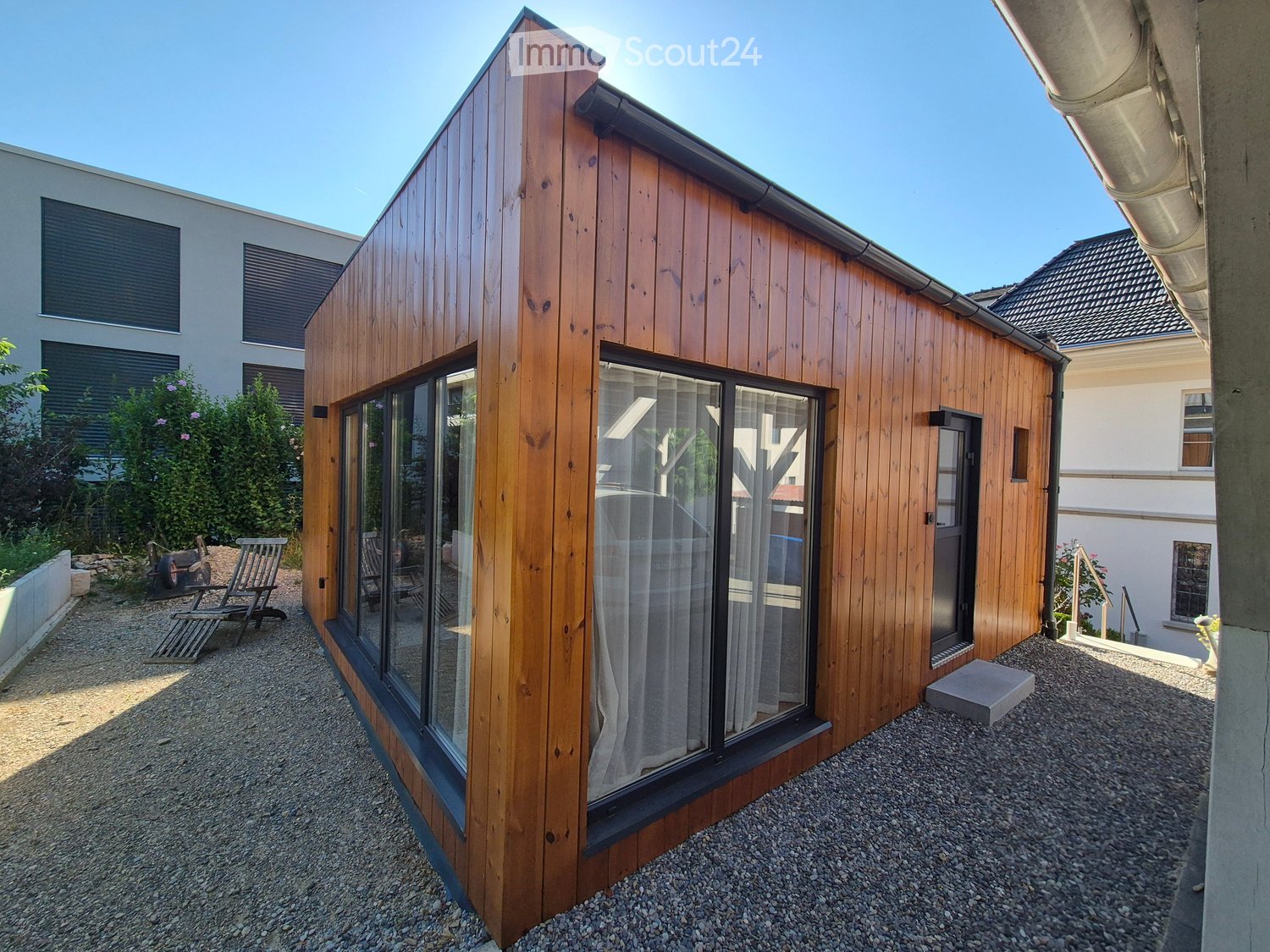 A modern wooden cabin with large glass windows and a gravel-covered ground area, surrounded by greenery and other buildings.