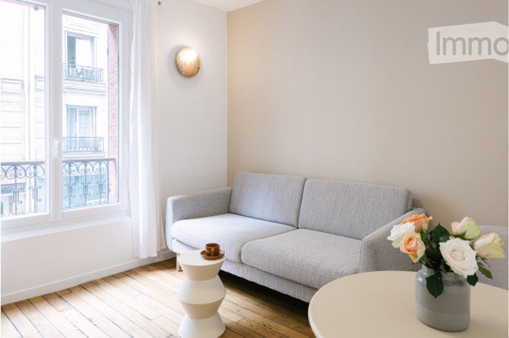 Modern living room with wooden floor, large window with view, grey sofa, two white stools, round table with vase of flowers.