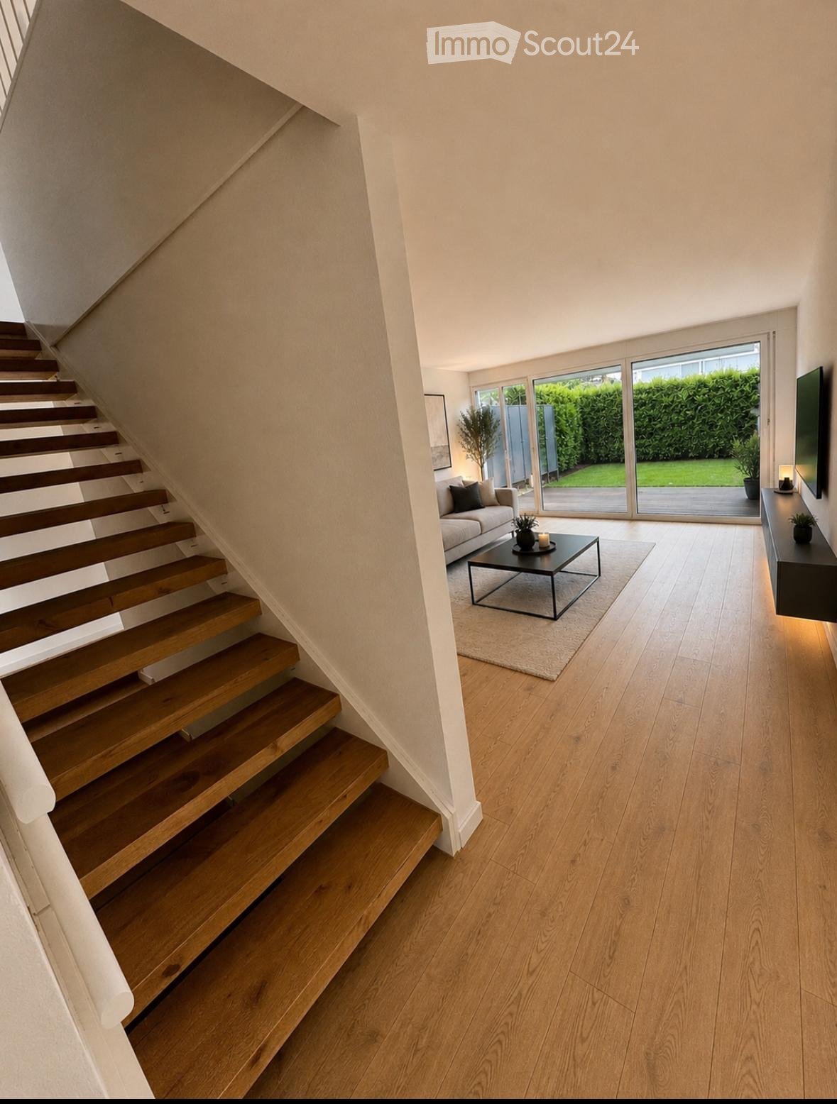 Modern living room with wooden flooring, a staircase leading upwards, a sofa, coffee table, and a TV screen. Large windows provide natural light and a view of the garden.