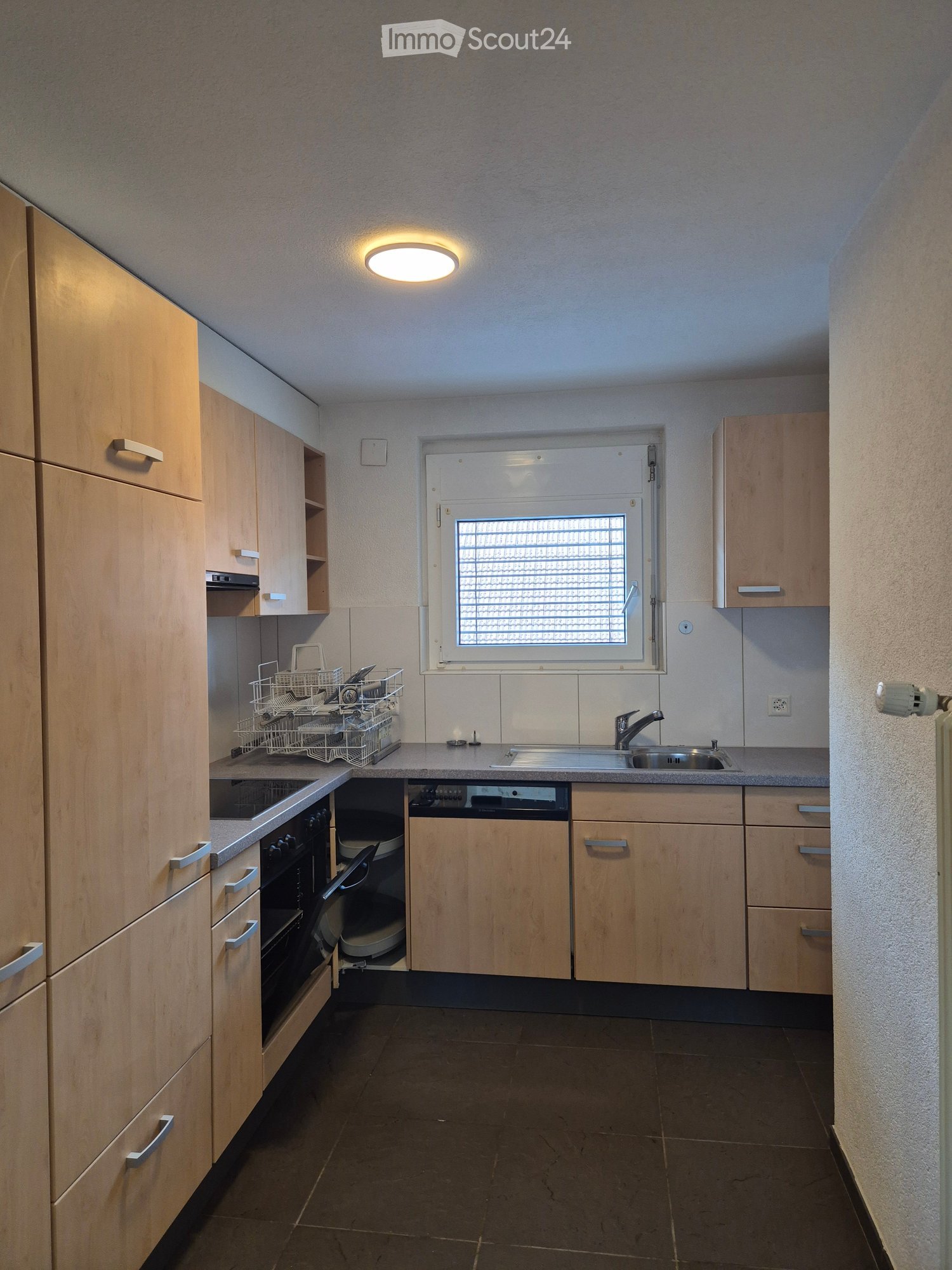 Modern kitchen with light wood cabinets, stainless steel appliances, and a window with blinds. Includes a dishwasher, oven, sink, and dish rack.