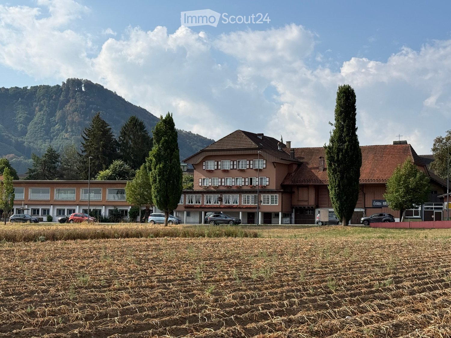 Two-story building with red-tiled roof and multiple windows, surrounded by trees and parked cars, with a grassy field in the foreground and mountains in the background.