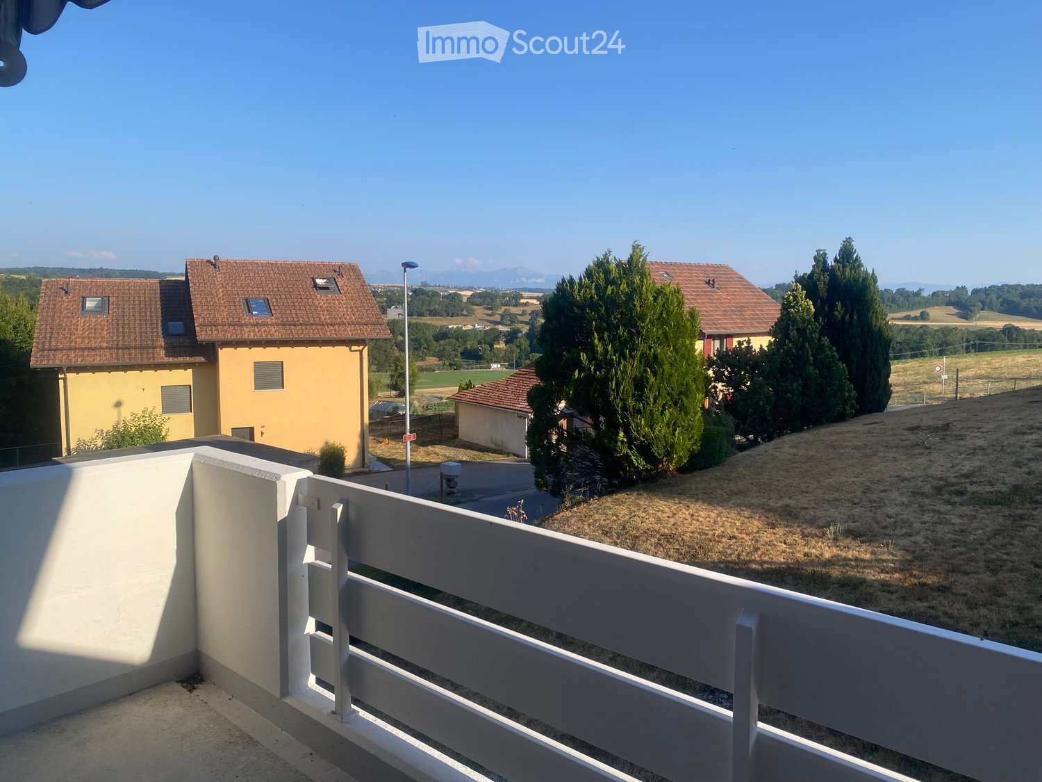 Balcony with white railing overlooking a rural landscape with houses, trees, and open fields.