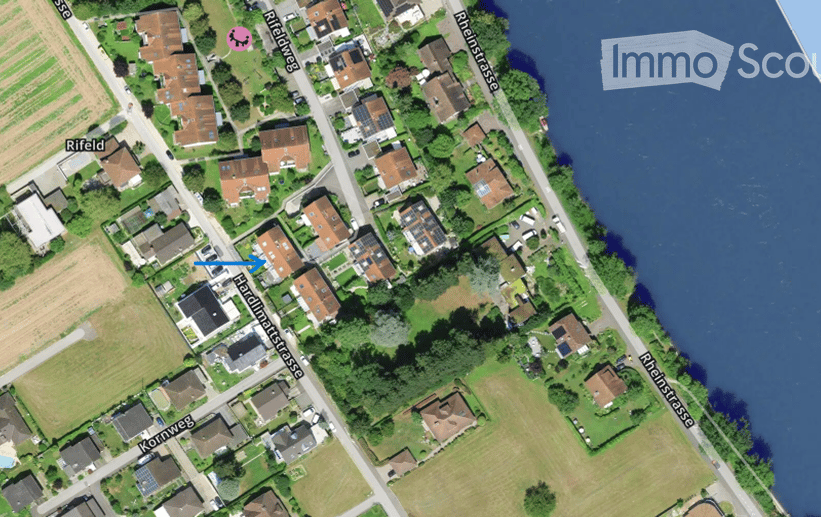 Aerial view of a residential area with multiple houses, gardens, and surrounding fields. The houses are clustered along streets, with some near a body of water.