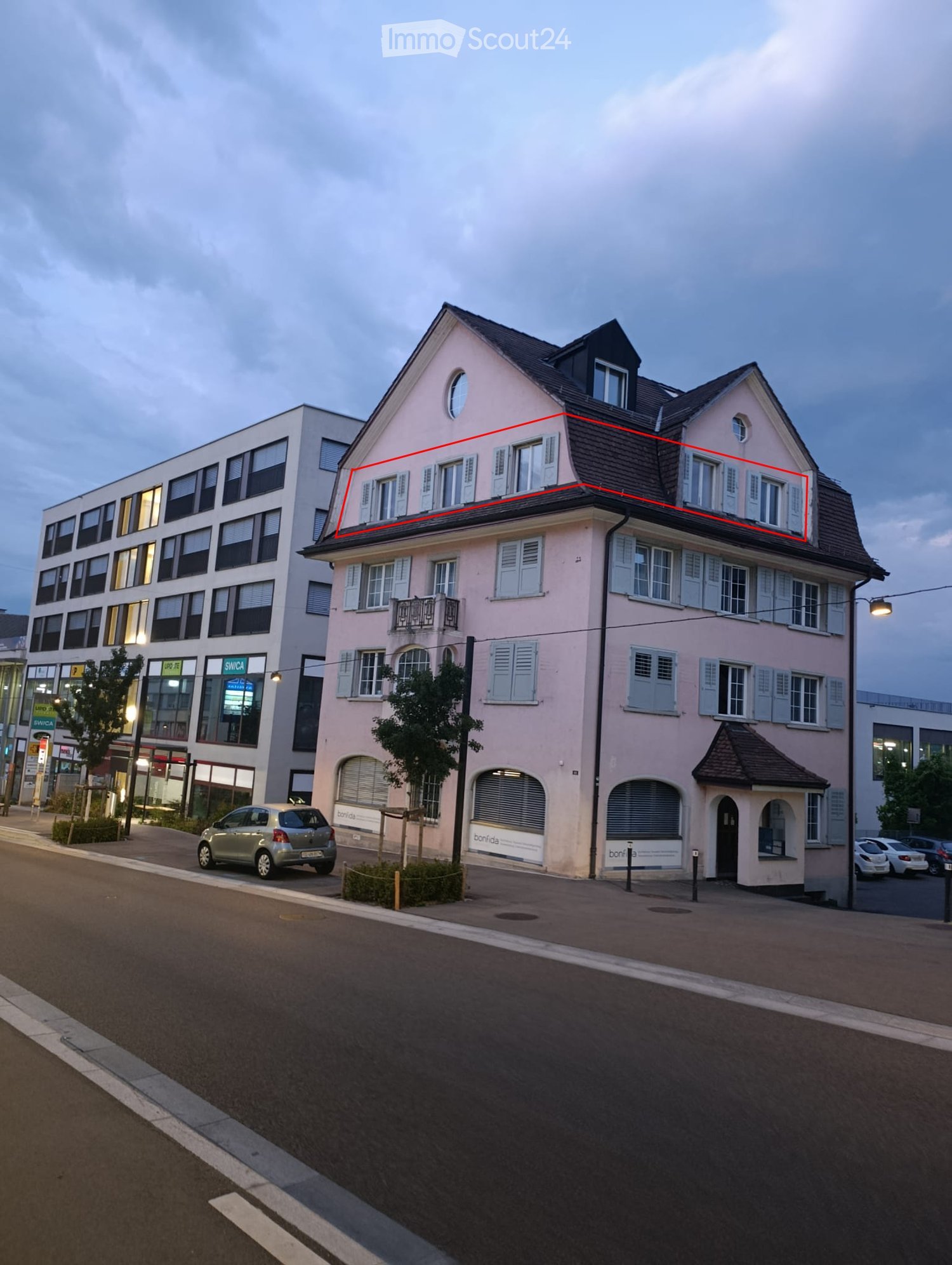 A two-story residential building with a pink facade, multiple windows, and a small balcony on the upper floor. The building is situated on a street with a parked car and some greenery nearby.