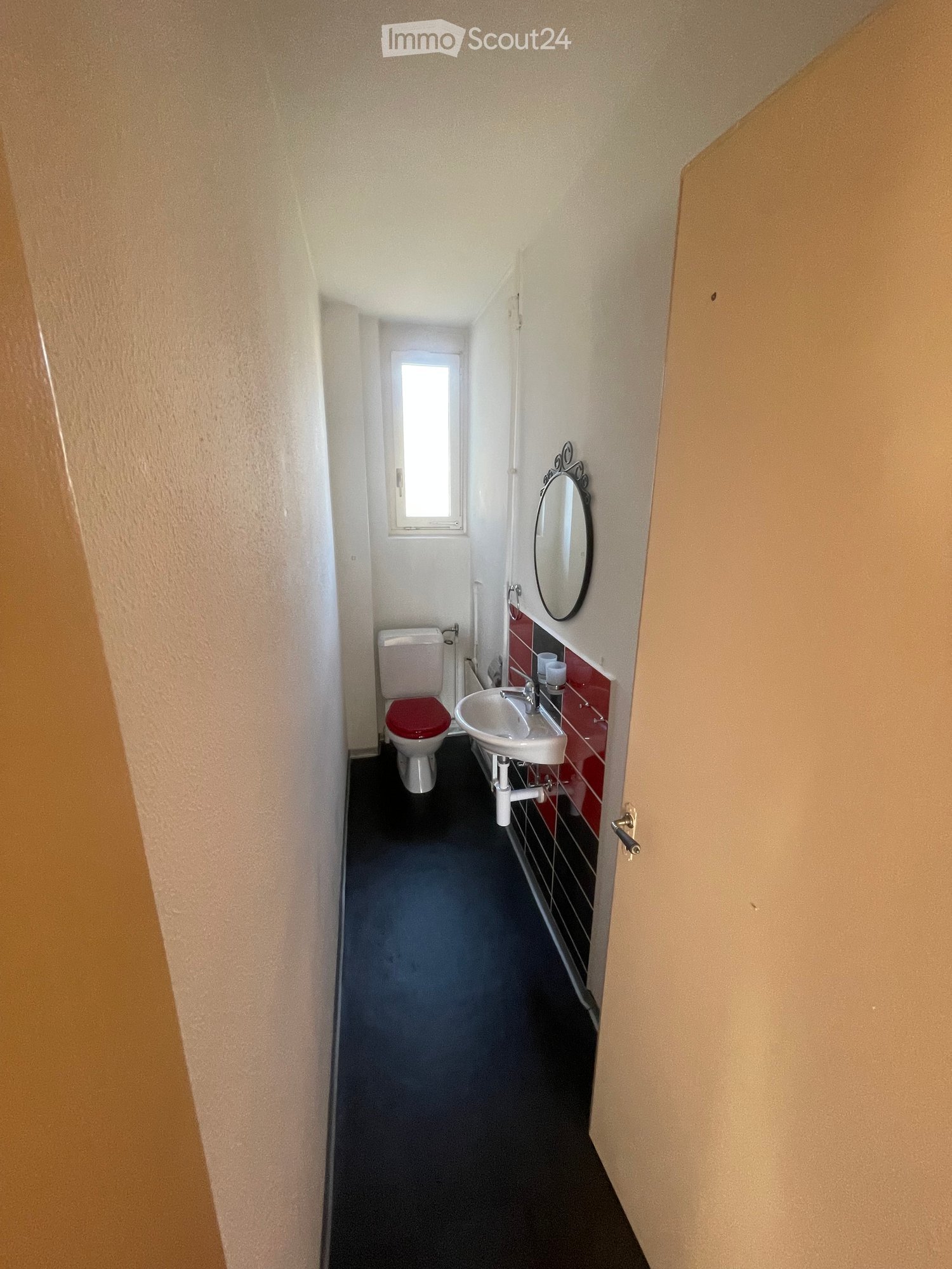 Small bathroom with a toilet, sink, and mirror. The walls are painted in a light color with red tiles around the sink area. The floor is dark grey.