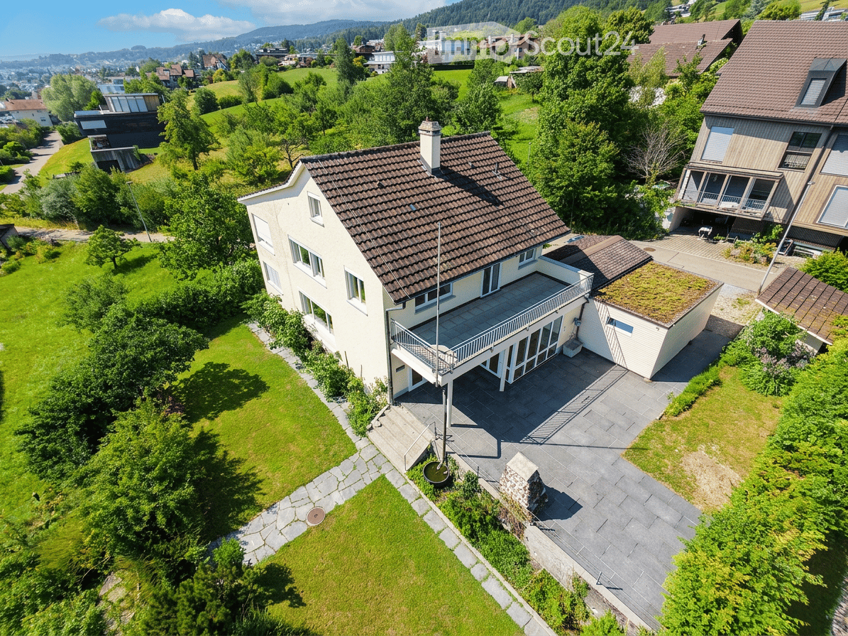Aerial view of a two-story house with a brown tiled roof, white walls, and a spacious balcony. The property is surrounded by a well-maintained lawn and trees, with neighboring houses visible in the background.