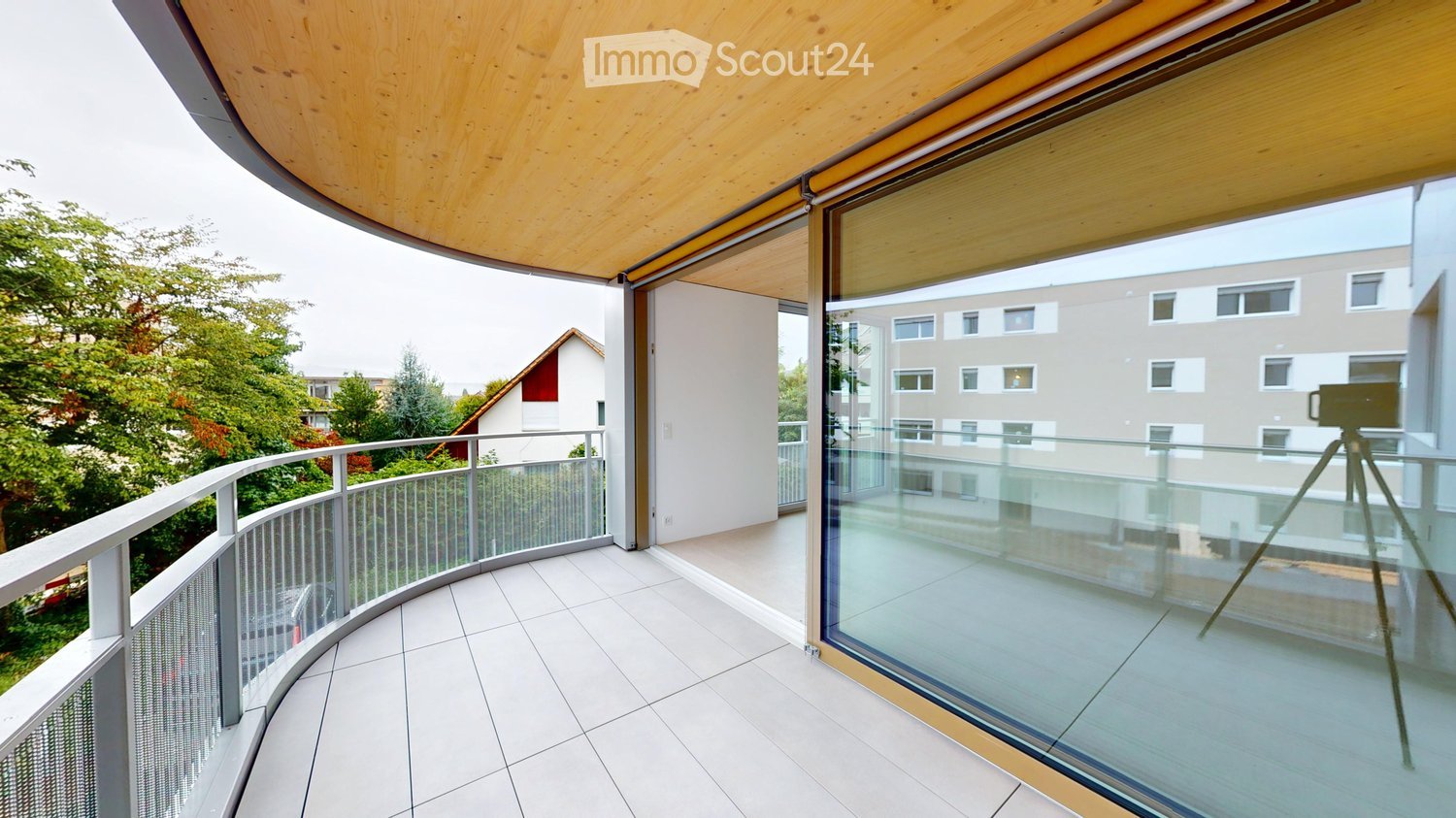 Modern balcony with white tiled floor, curved glass railing, and wooden ceiling. Offers views of greenery and neighboring buildings.
