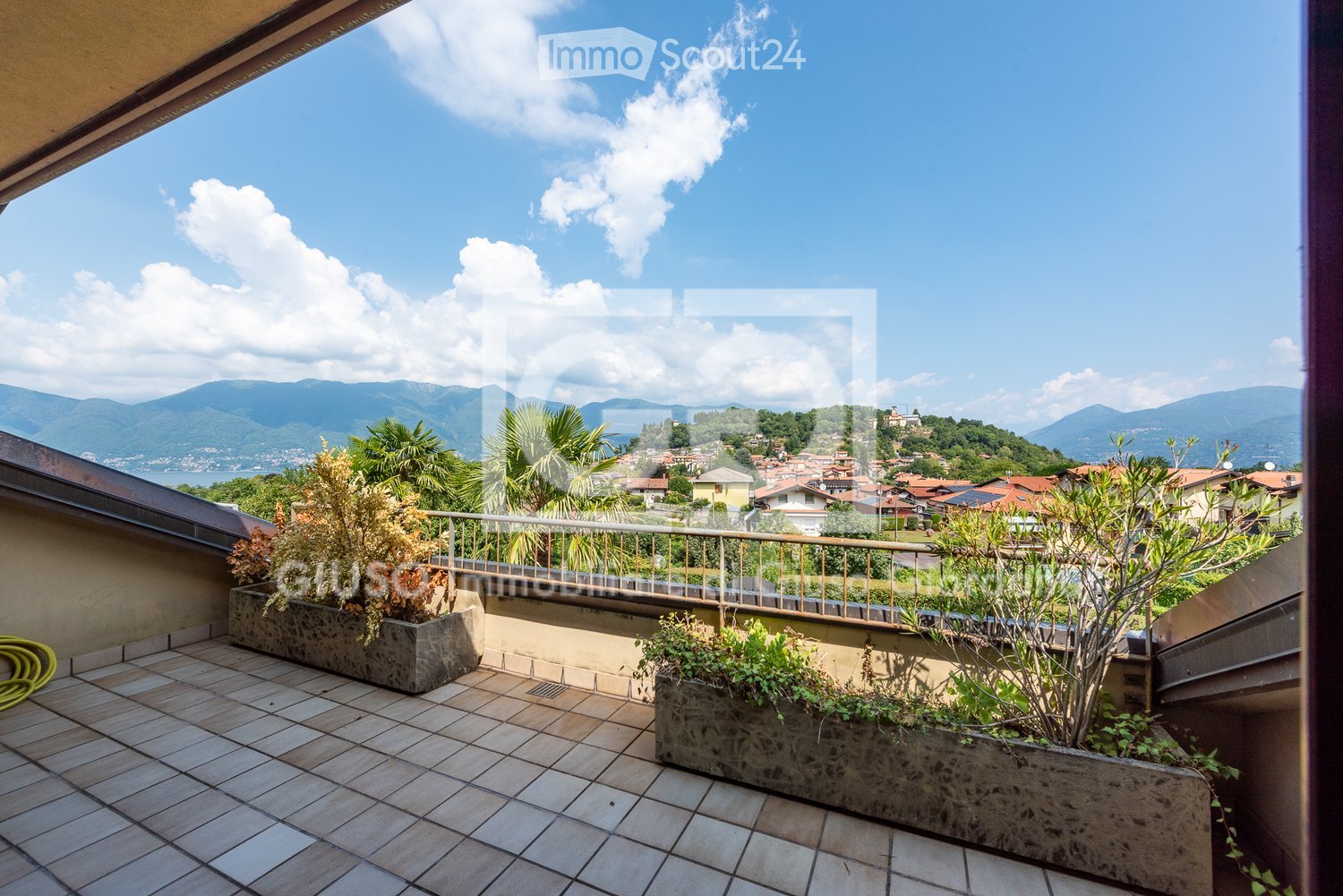 Balcony with tiled flooring, metal railing, and potted plants, overlooking a scenic town and mountainous landscape.