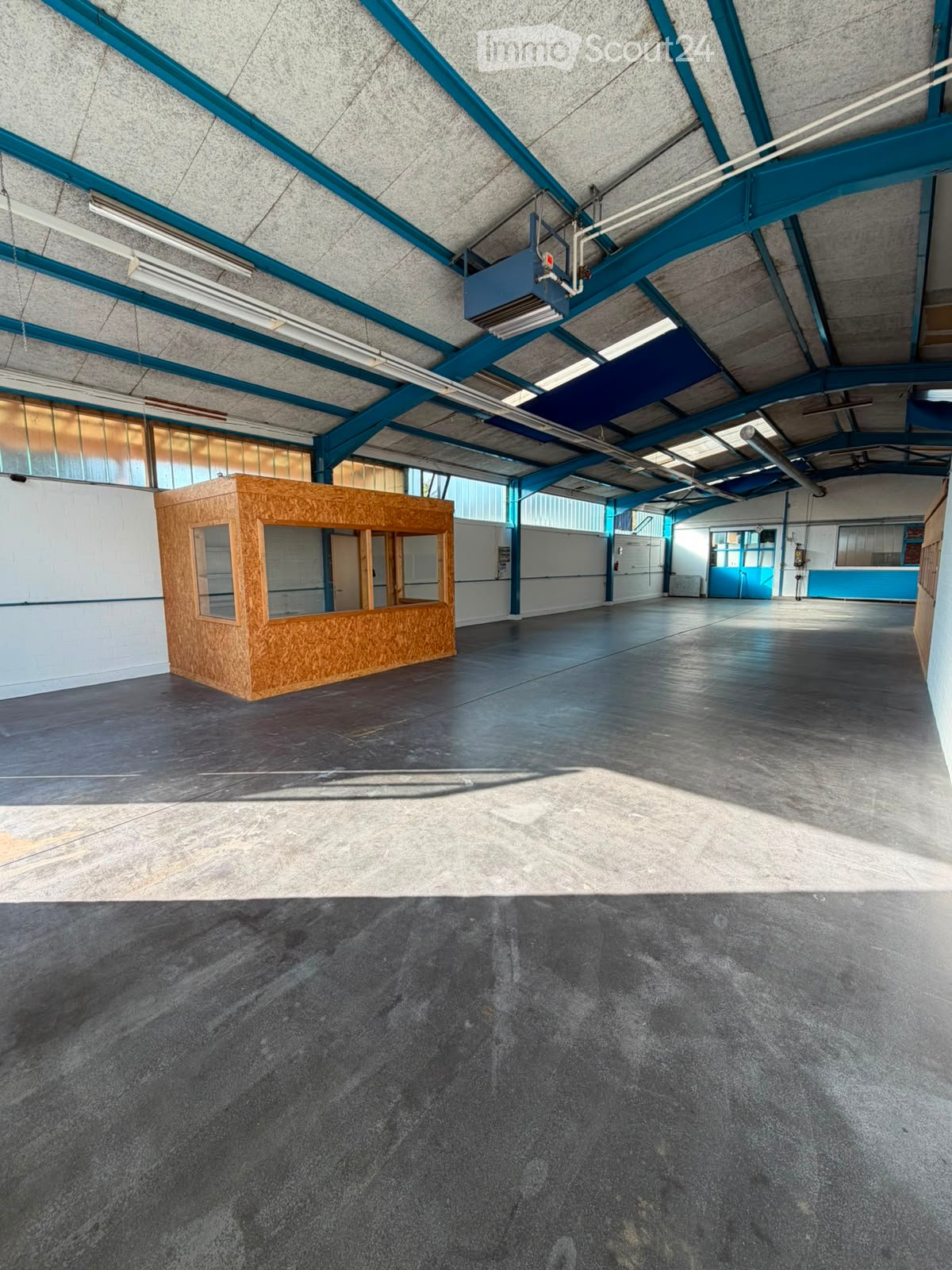 Large, open industrial-style space with a concrete floor, exposed steel beams, and a small wooden booth. The area is well-lit with natural light from large windows and artificial lighting from overhead fixtures.
