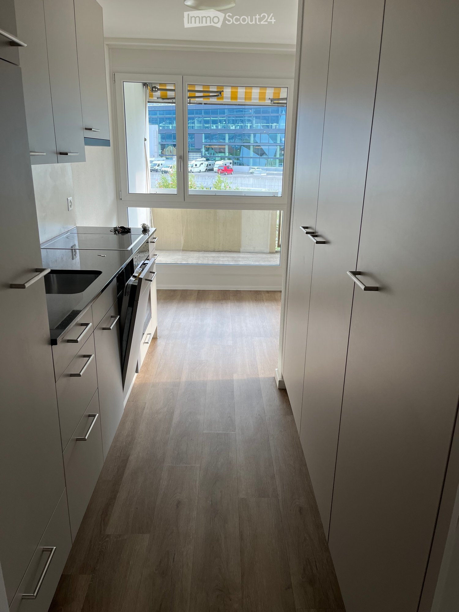 Modern hallway with light wooden flooring, white walls, and a large window at the end leading to an outdoor area. The hallway features sleek cabinetry on one side and a glass door with blinds.