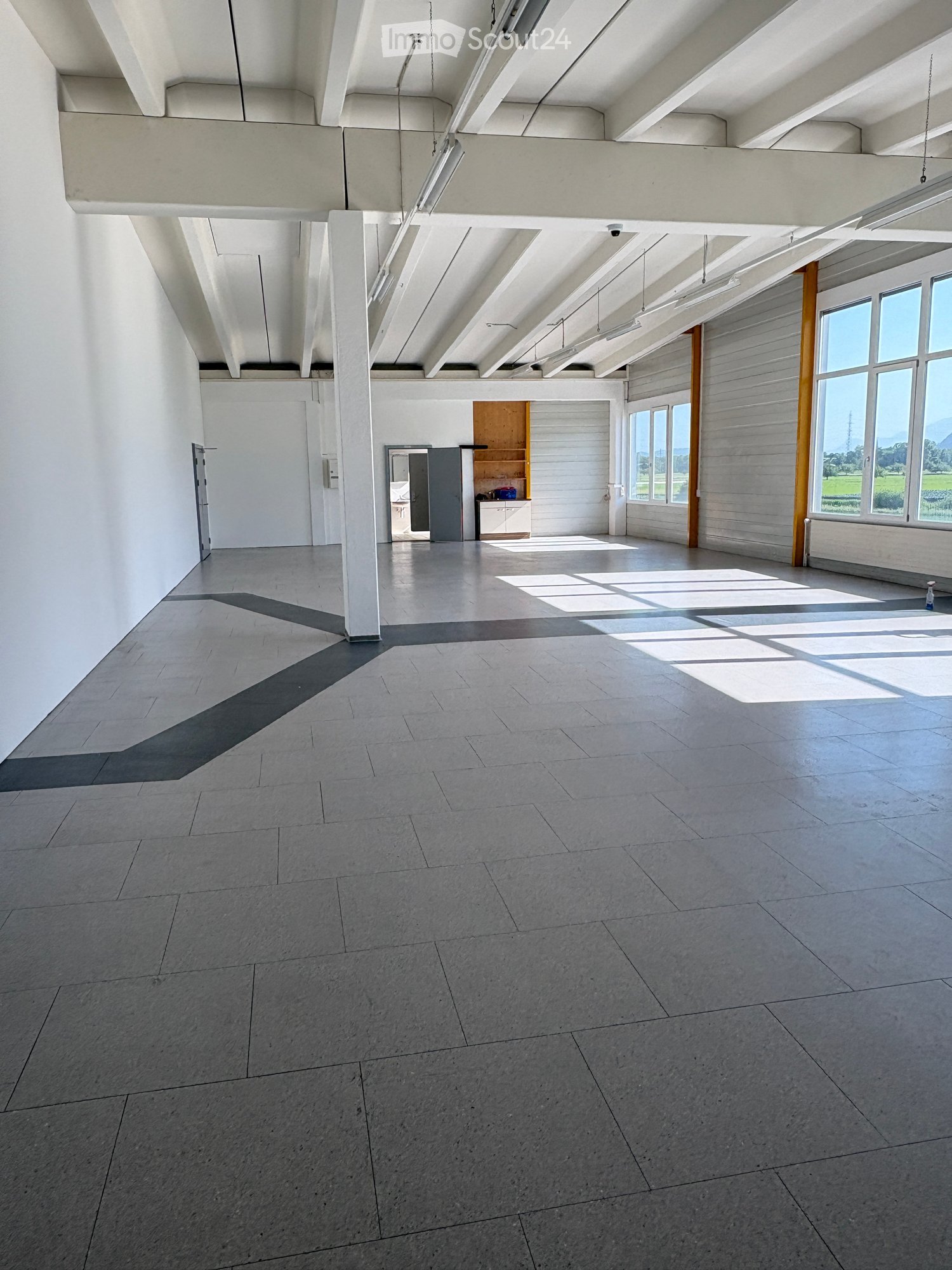 Empty room with white walls, concrete floor, large windows, and a small cabinet in the background.