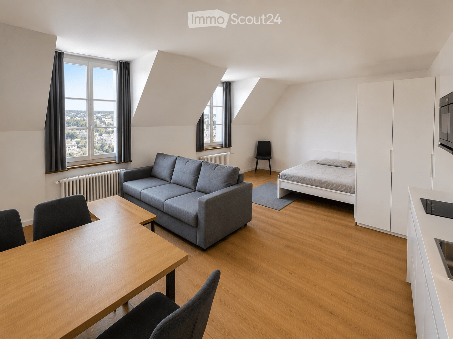 A modern, compact studio apartment with wooden flooring, large windows offering a city view, a grey sofa, a wooden dining table with chairs, a single bed, and a small kitchen area with a microwave.