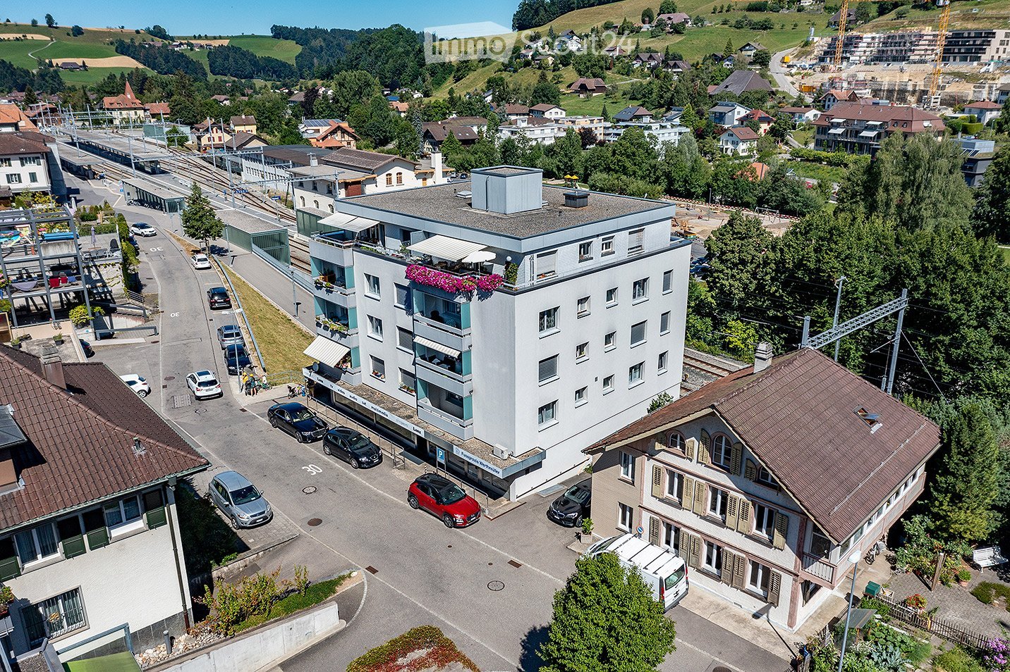 Aerial view of a modern multi-story building with balconies and a rooftop terrace, surrounded by parked cars and other residential structures in a suburban area.