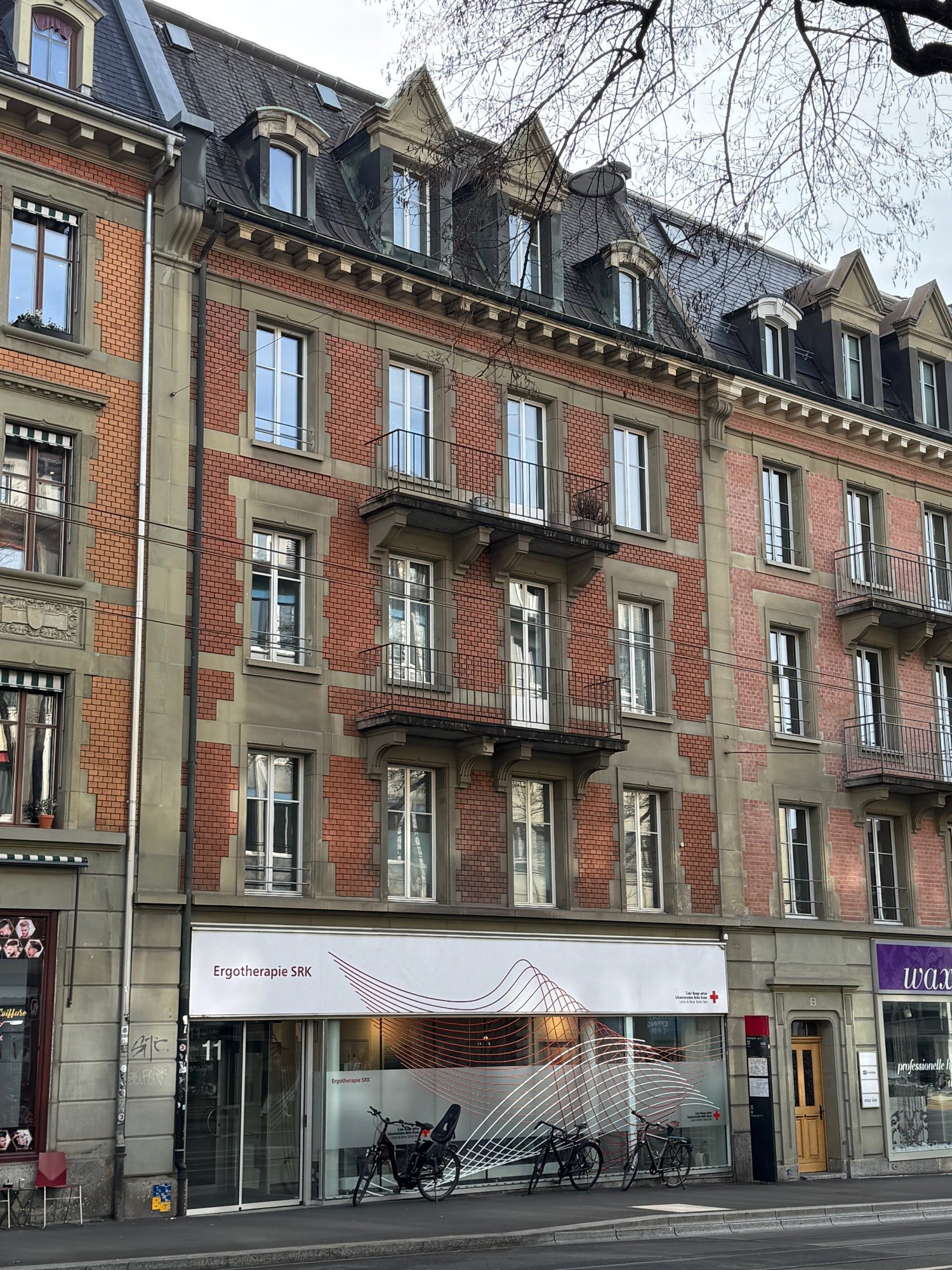 A multi-story building with brick facade, multiple windows, and balconies. The ground floor has a glass storefront with a sign reading Ergotherapie SRK.