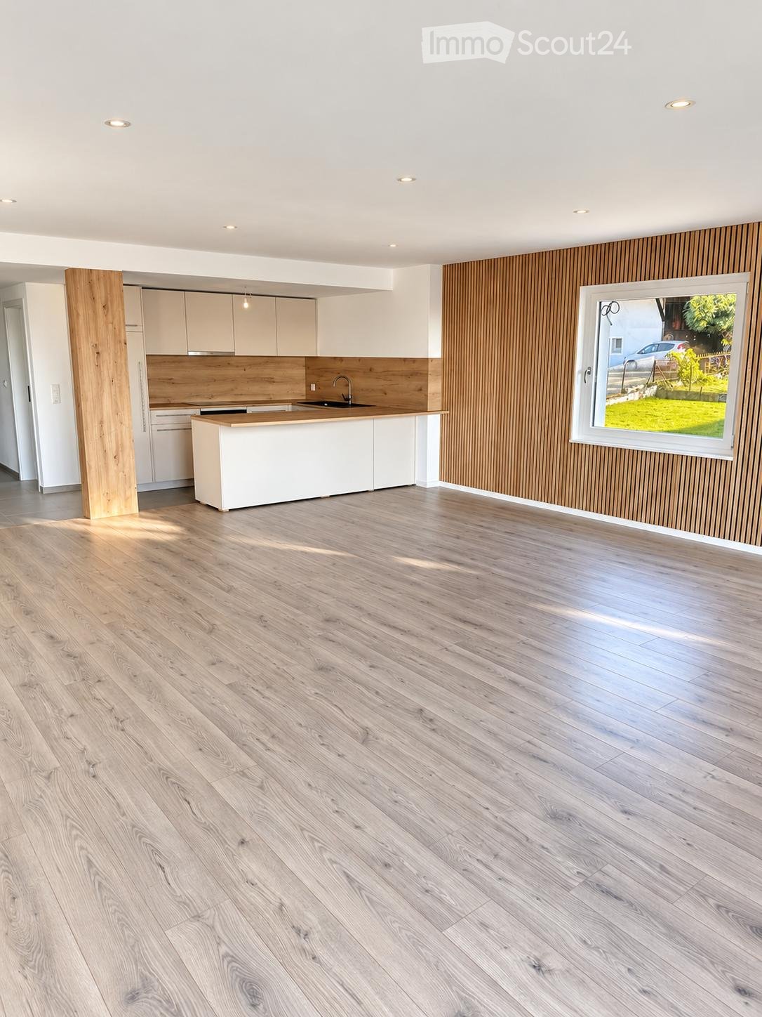 Empty kitchen with light wooden flooring, white cabinets, and a wooden backsplash. A large window offers a view of the garden and a parked car.