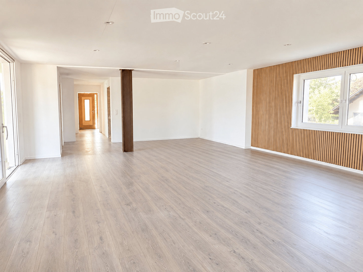 Empty room with light wooden flooring, white walls, and a large window with white frames. The room features a modern design with recessed ceiling lights and a wooden accent wall.
