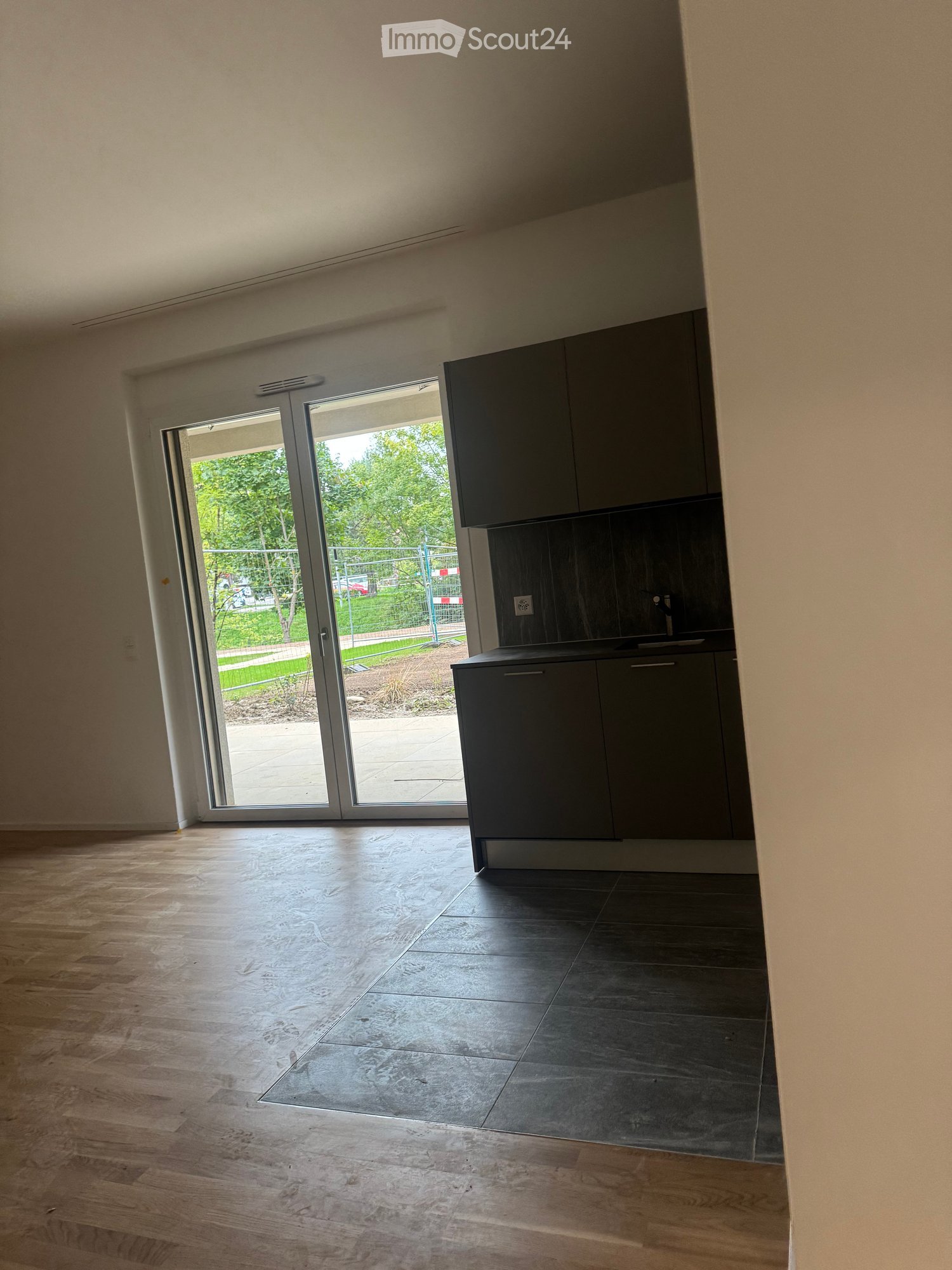 Empty room with light wooden flooring and dark tiled section, featuring a sliding glass door leading to an outdoor area with greenery visible.
