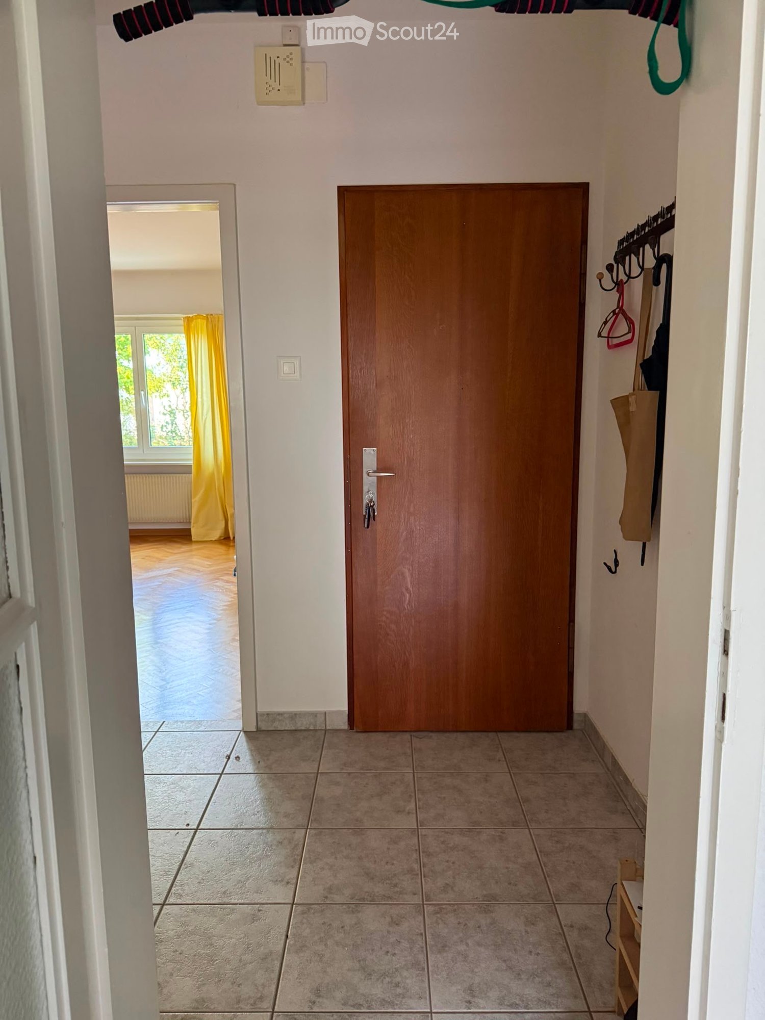A clean, tiled hallway with a wooden door, coat rack, and a glimpse of a room with yellow curtains and wooden flooring.