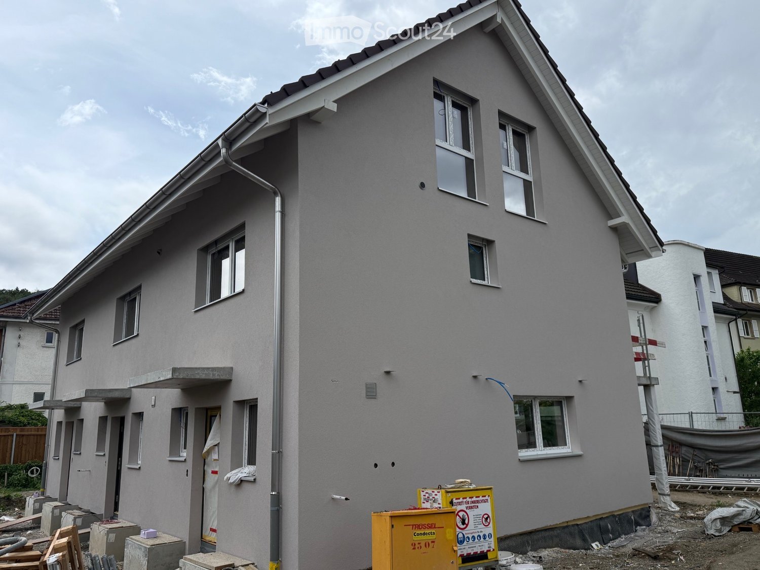 Two-story house with grey exterior, multiple windows, and a raised ground floor. The house is under construction with visible scaffolding and construction materials.