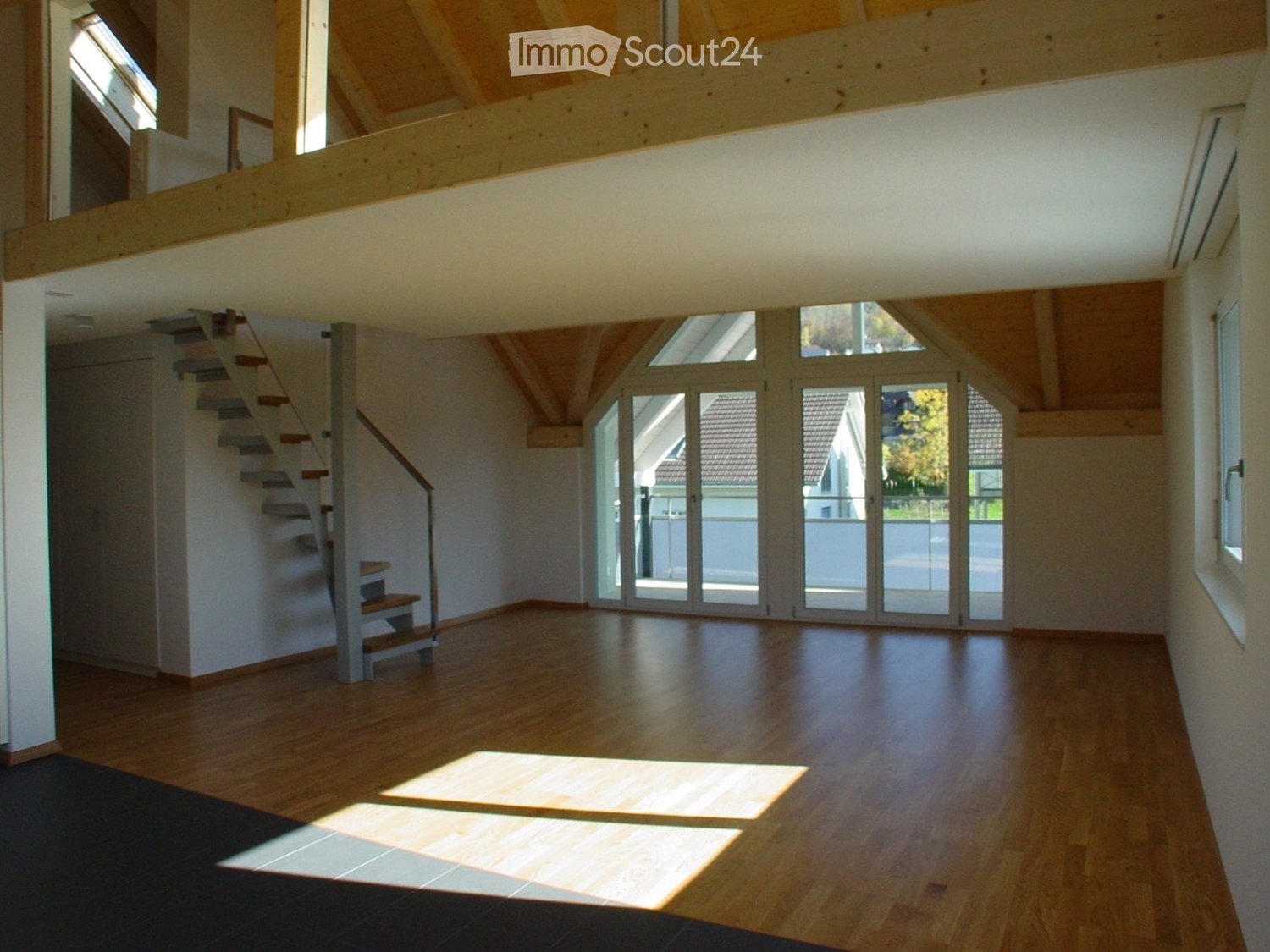 Bright, open living space with wooden floors, large windows, and a staircase leading to an upper level. The room features exposed wooden beams on the ceiling and ample natural light.