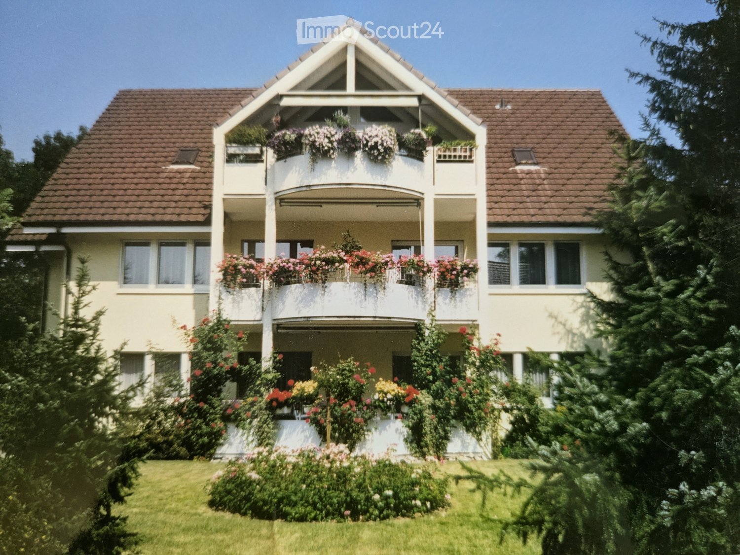 Two-story house with a brown tiled roof, white exterior, multiple windows, and balconies adorned with flower boxes and greenery.