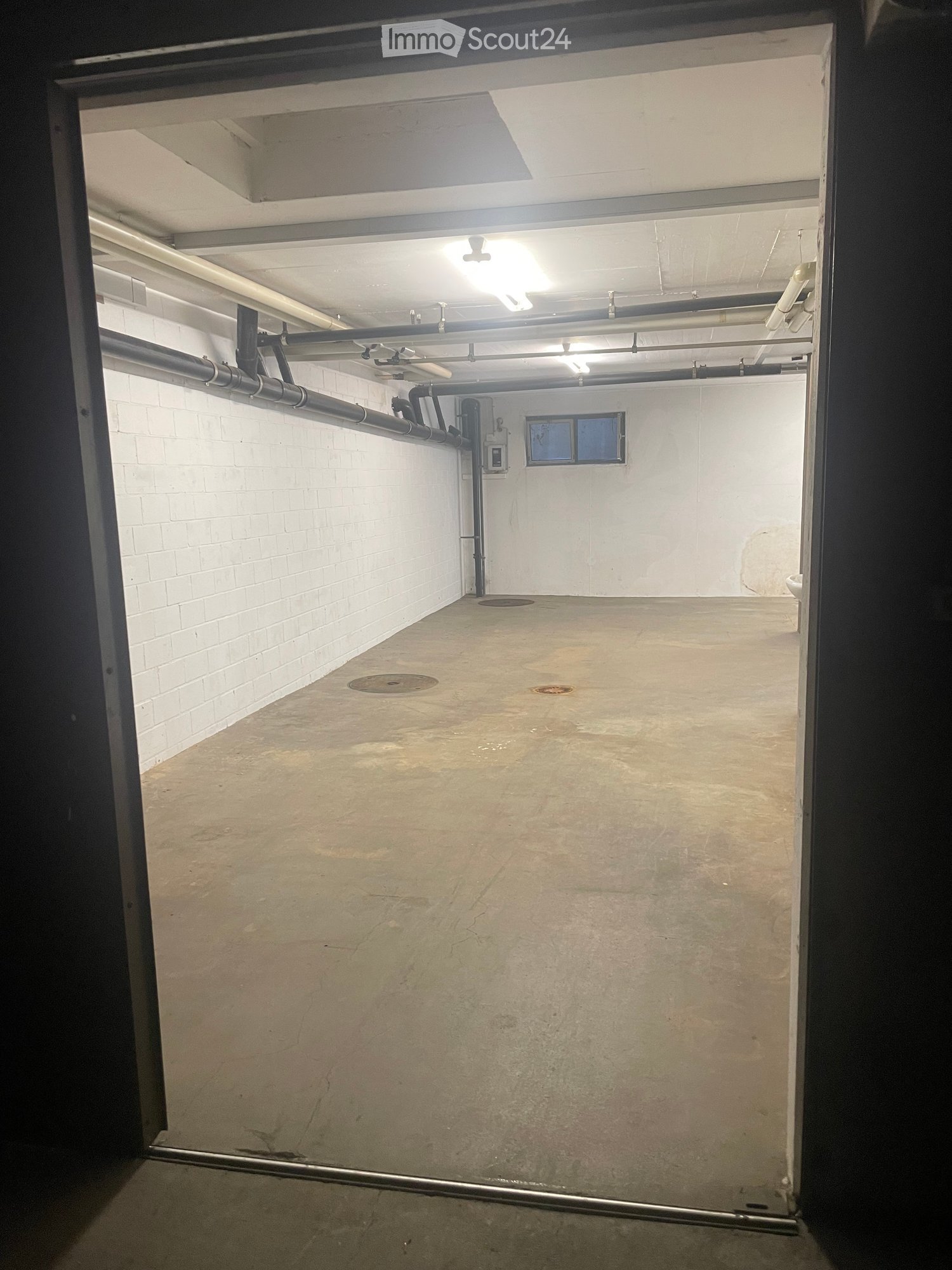 Empty basement or utility room with concrete floor, white brick walls, exposed pipes, and a small window at the far end.