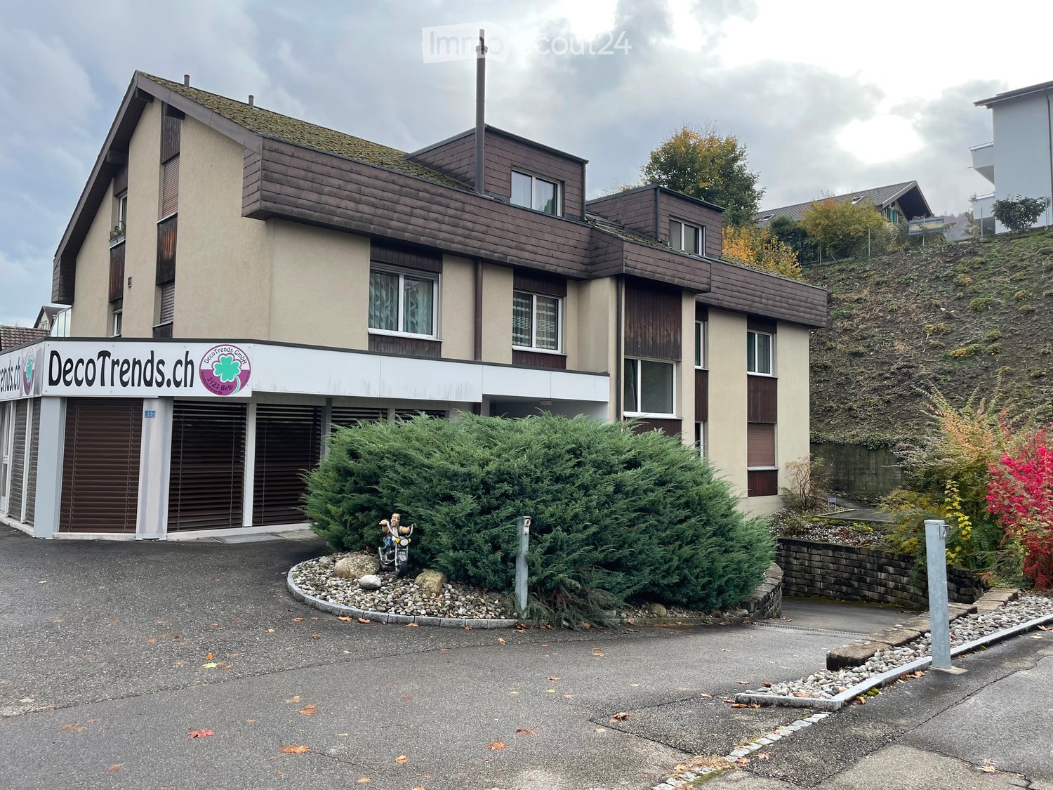 Two-story building with a shop front labeled "DecoTrends.ch" and a small garden area with bushes and a decorative statue.