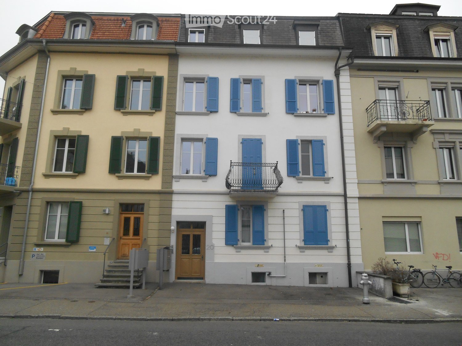 Three-story residential building with multiple windows, shutters, and balconies. The central building has a white facade with blue shutters and a small balcony. The left building has a yellow facade with green shutters, and the right building has a light