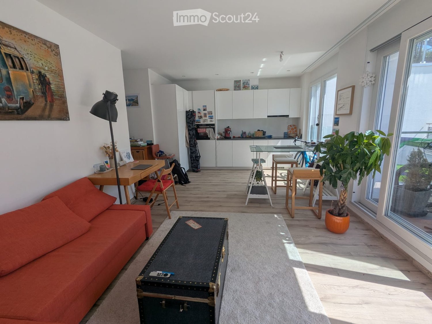Open-plan living and dining area with a red couch, coffee table, and a dining table with chairs. A bicycle is parked near the kitchen. A potted plant and a lamp are on the floor. The kitchen has white cabinets and a microwave.
