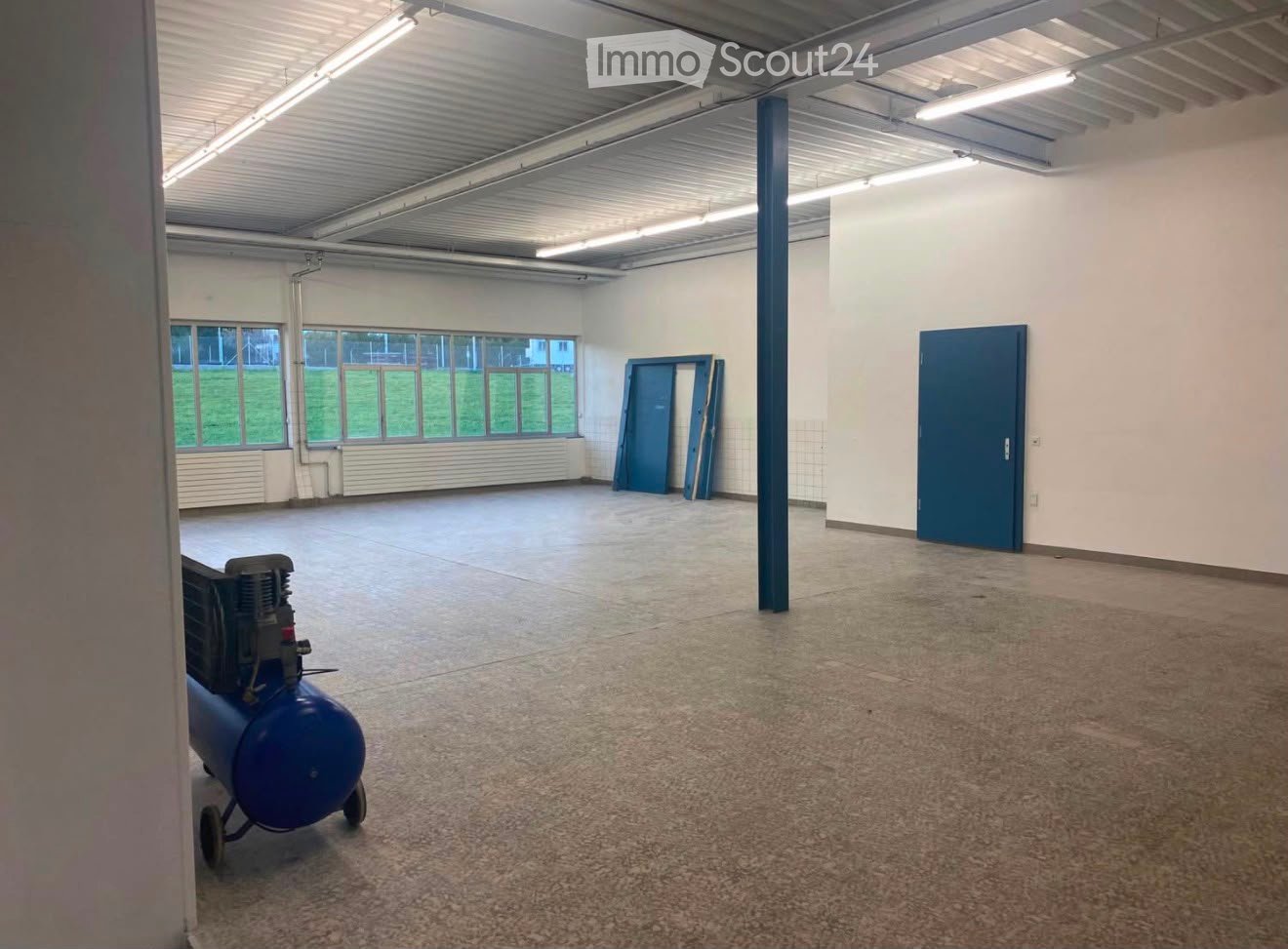 Empty room with bare walls, concrete floor, large windows, and a blue air compressor in the corner.