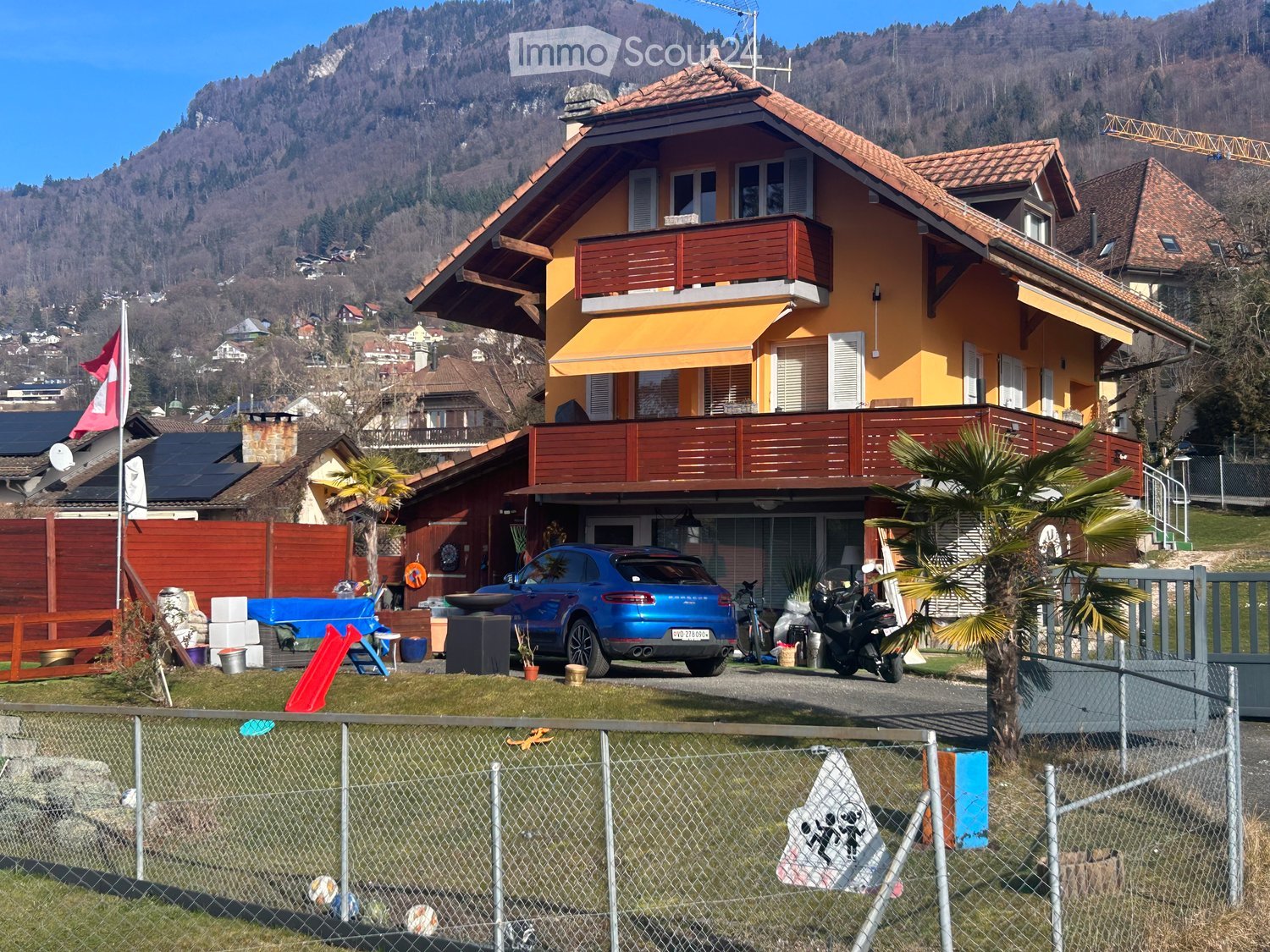 Two-story yellow house with red wooden balconies, surrounded by a fenced yard with a blue car, motorcycle, and various outdoor items.