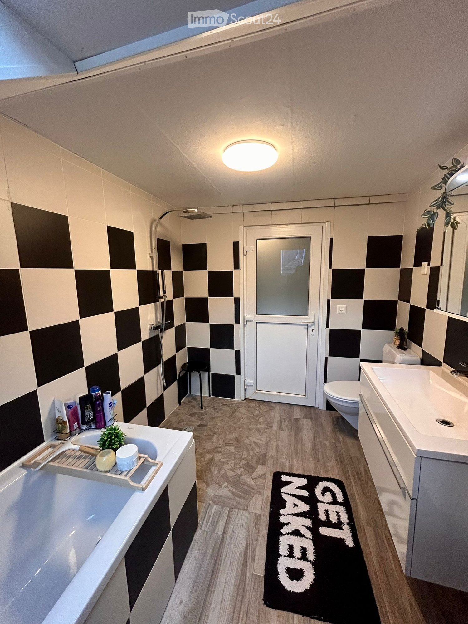 Modern bathroom with checkered black and white tiles, wooden flooring, bathtub, sink, toilet, shower, and a small stool. Includes a black bath mat with white text and a small plant on the sink.