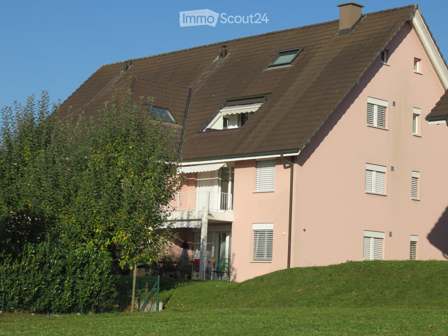 Two-story pink house with a brown tiled roof, multiple windows with shutters, a small balcony, and a raised ground floor.