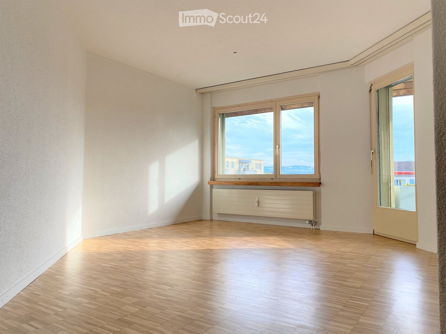 Empty room with wooden flooring, white walls, and large windows offering a view of the sea and buildings.
