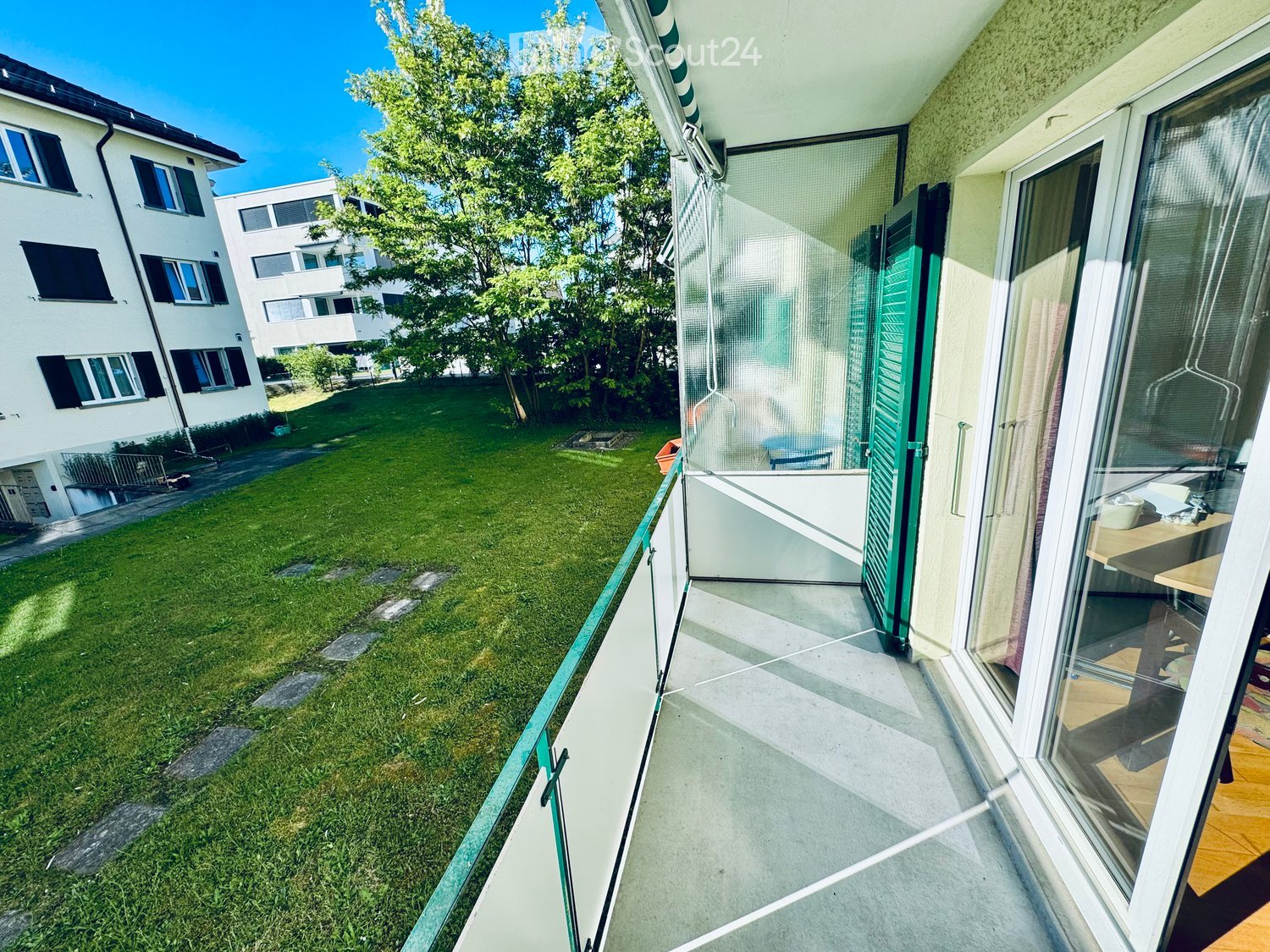 Balcony with a view of a grassy yard and neighboring buildings, featuring a glass door and a small table inside.