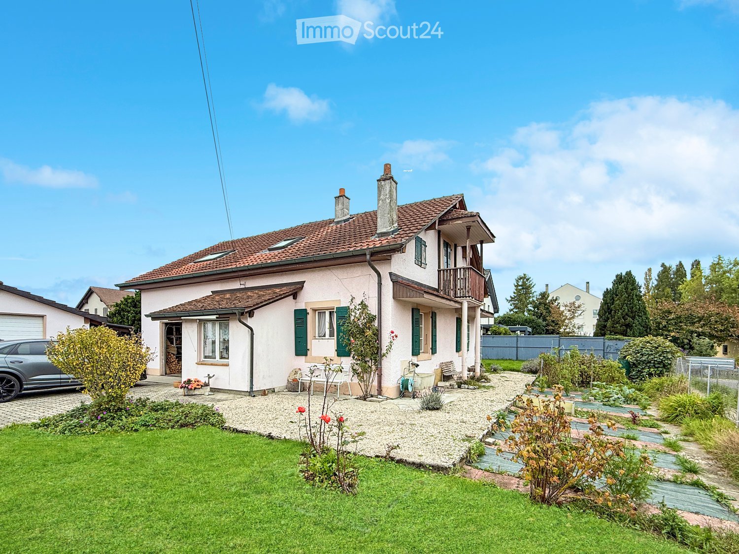 Two-story house with a tiled roof, green shutters, and a balcony. The front yard features a well-maintained lawn, garden beds, and a small patio area with outdoor furniture.