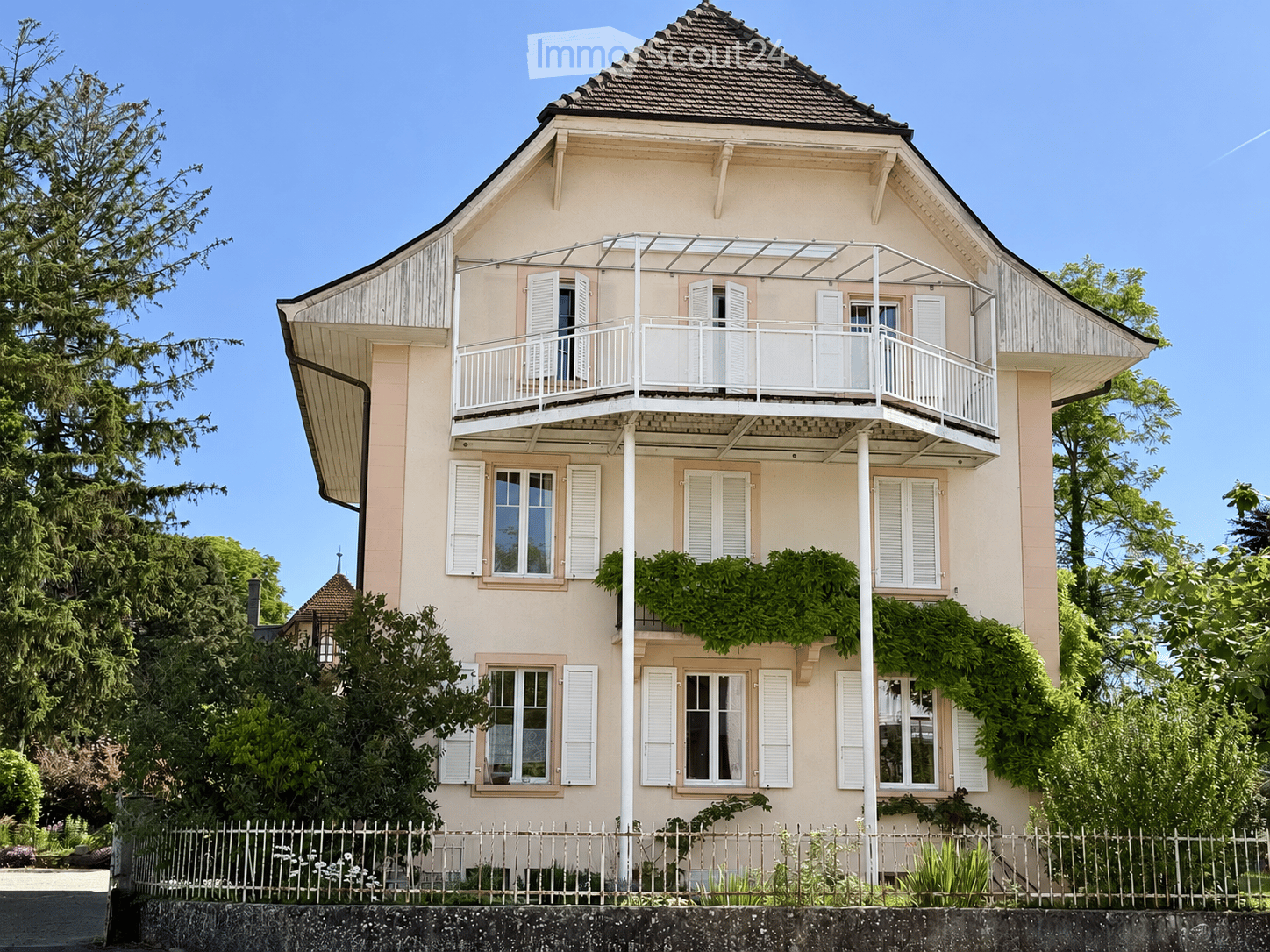 Two-story house with light pink facade, white shutters, and a balcony on the upper floor. The house has a sloped roof and is surrounded by greenery and a white fence.