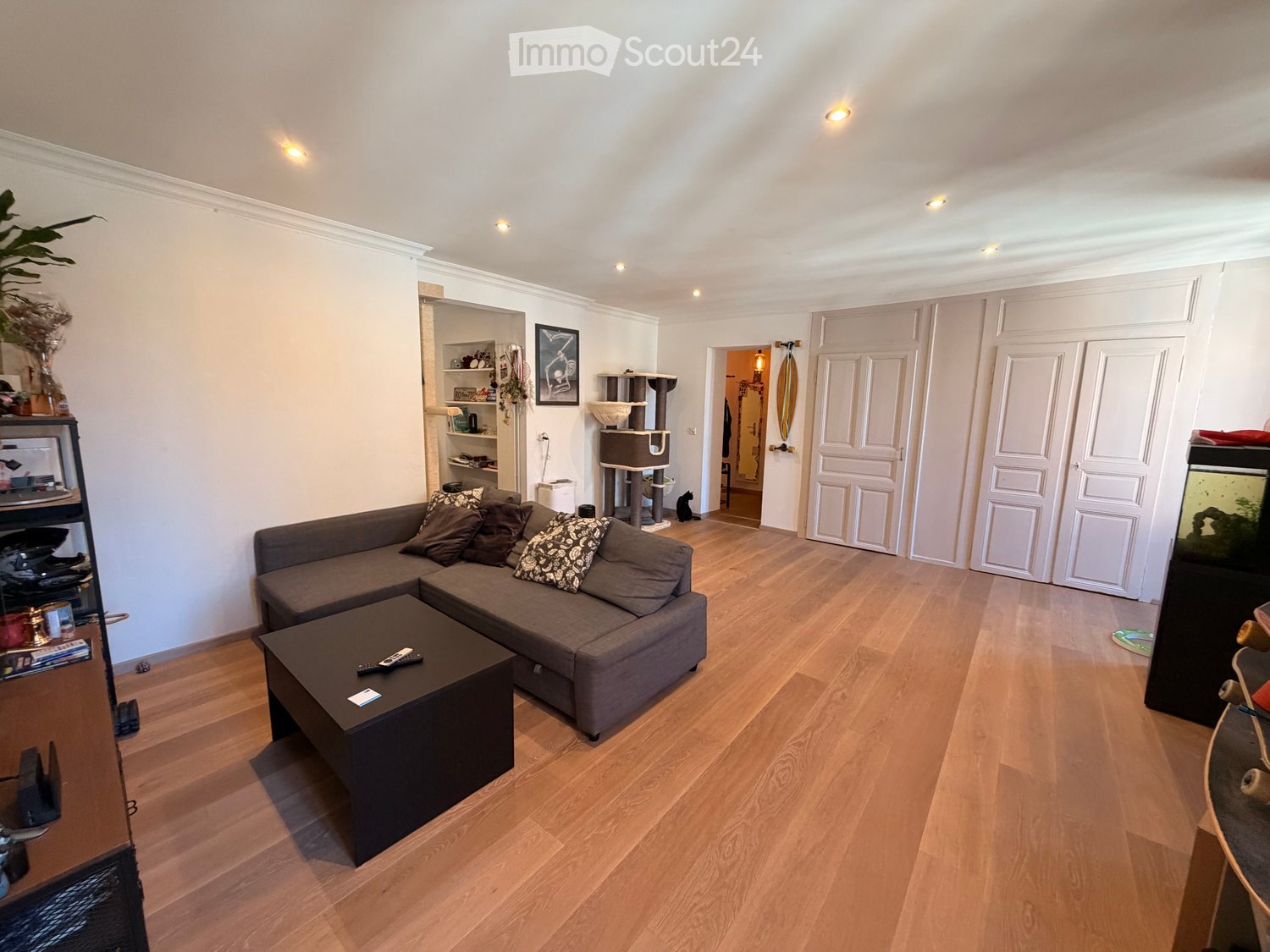 Modern living room with light wooden flooring, grey sectional sofa, coffee table, cat tree, shelves, and fish tank.